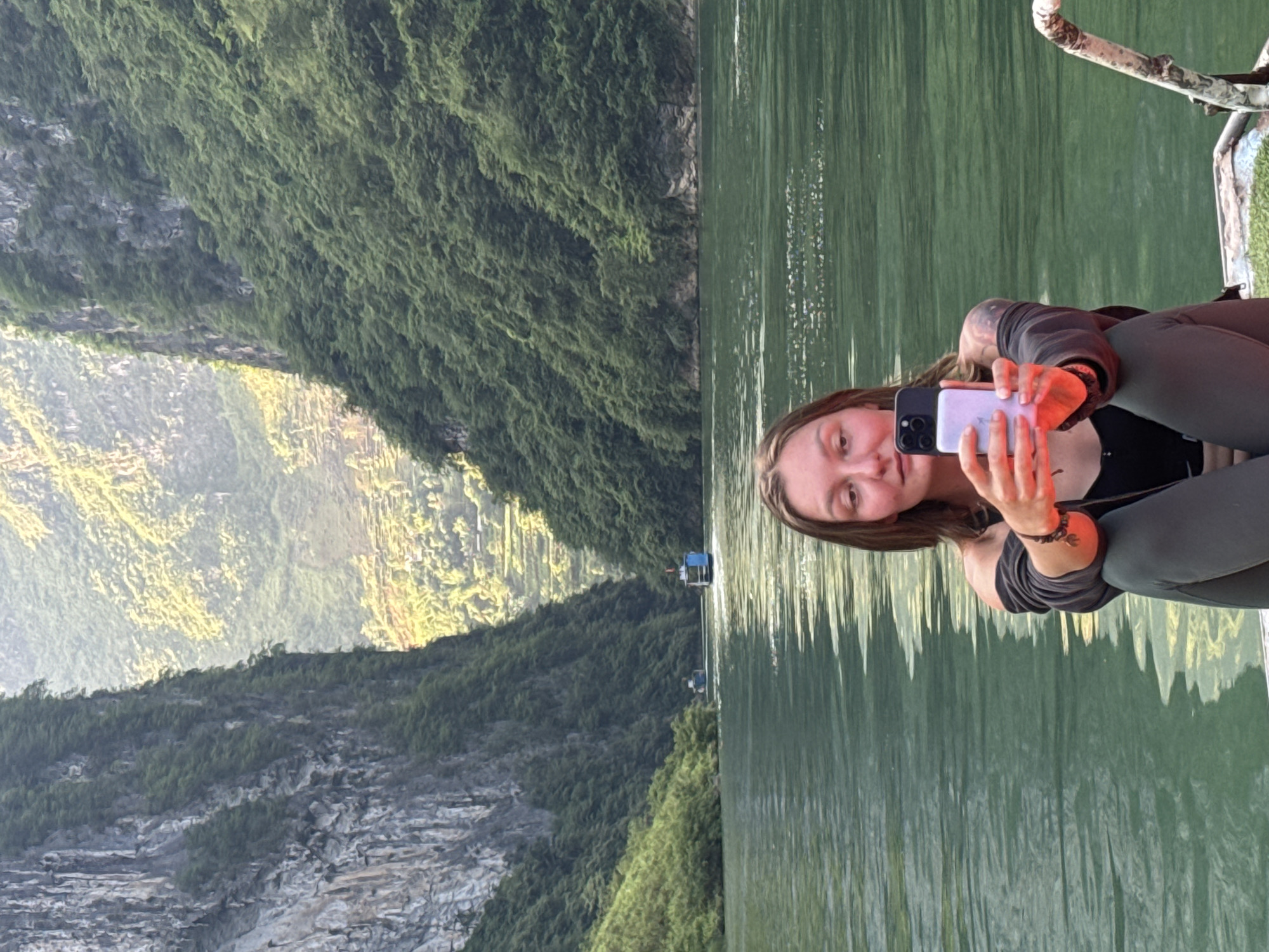 A young woman is holding up a smartphone and taking a selfie while sitting on a boat or dock on a body of water. The water is calm with some surface reflections. Behind her is a mountainous landscape, partly covered in dense green vegetation and some rocky areas. There appear to be some structures or buildings along the shoreline. While it is hard to verify that the location is exactly the city of Đồng Văn Commune, Vietnam, the landscape is highly typical of the geography of the city.