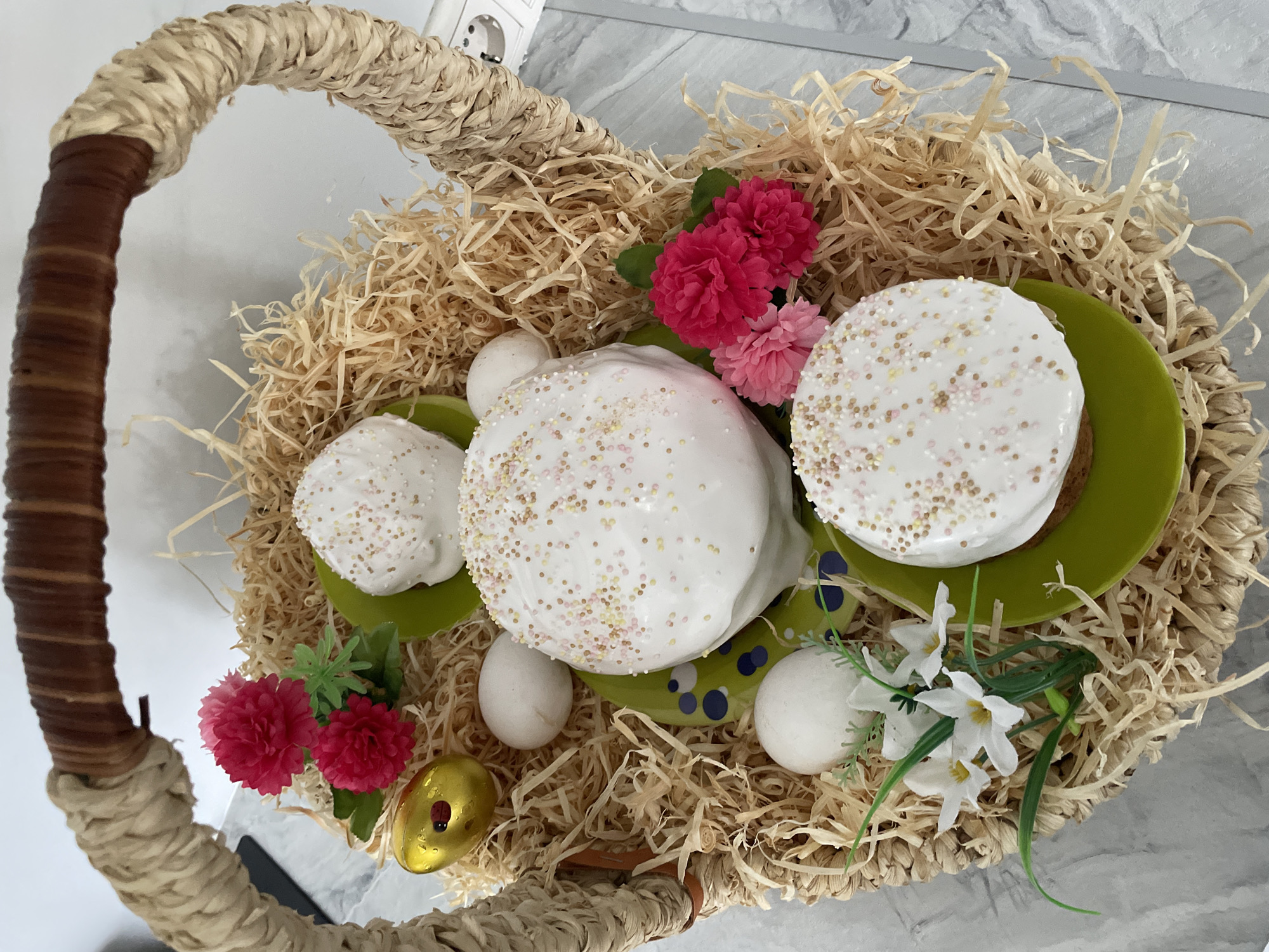 The image shows a wicker basket filled with straw-like material. Inside the basket are three decorated cakes, likely Easter cakes, topped with white icing and sprinkles. They sit on green plates. Also within the basket are three white eggs and one golden egg. Floral decorations, including pink and white flowers with green leaves, are scattered around the cakes and eggs. The handle of the basket is visible on the left and top of the image. The background is a light gray surface, possibly a countertop. The location of the image is Лесной Пейзаж Кп (шарапово, Russia.