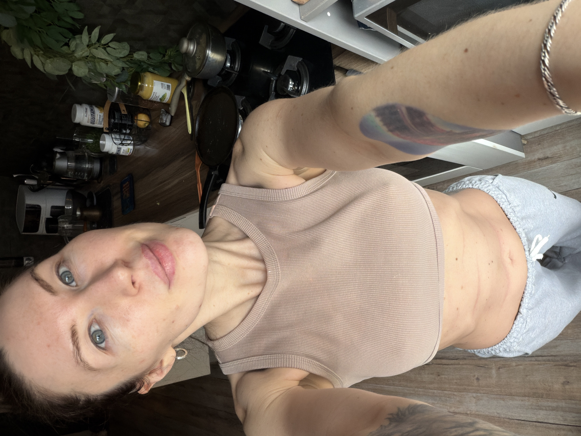 A woman is taking a selfie in a kitchen. She is wearing a beige ribbed tank top and grey sweatpants. The kitchen contains a stovetop, various kitchen utensils, and bottled goods. The location is Perm, Russia.