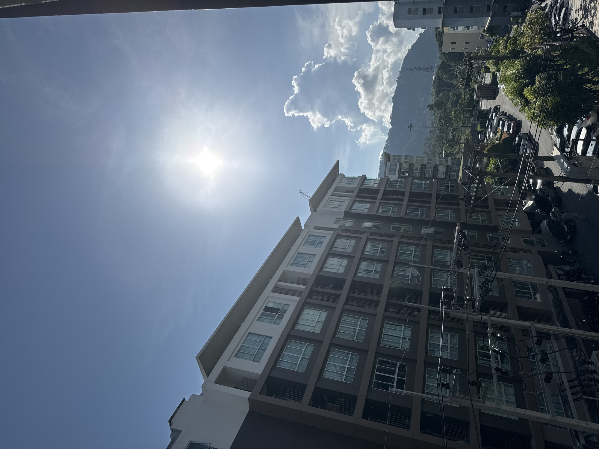 The image shows an exterior view from an elevated position, likely a balcony, looking towards a multi-story apartment building and a street below. The sky is bright blue with the sun prominently visible, casting a glare and a starburst effect. A large, fluffy white cloud is situated in the mid-right portion of the sky, above a lush, green, mountainous landscape in the background.
The apartment building in the foreground is modern, with a dark brown facade on the lower floors and white sections on the upper floors. It features numerous windows with light-colored curtains. Balconies with dark railings are visible between floors. In front of the building and extending towards the street are numerous utility poles with a complex network of wires and electrical components.
Below the apartment building, a paved street is visible, lined with parked vehicles including cars and motorcycles. Some trees and greenery are present along the street and on the slopes of the hills in the background. The scene suggests an urban or semi-urban environment with a tropical climate, consistent with the location of Kathu, Thailand. No people are clearly identifiable in the image. The overall impression is one of a sunny day in a residential area.