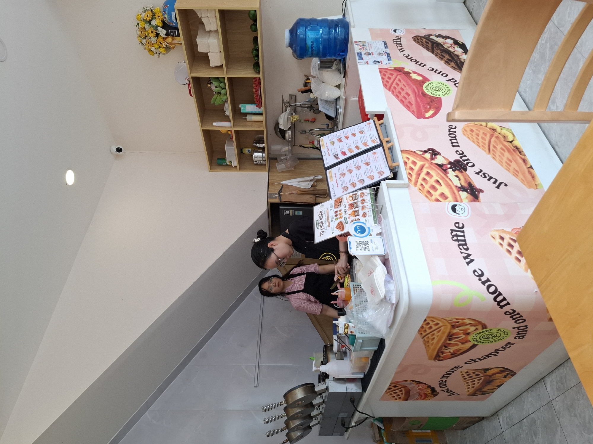 The image depicts the interior of a waffle shop located in Nha Trang, Vietnam. Two young women are working behind a counter. One woman, wearing a black apron over a black shirt, is actively preparing food, holding a knife and looking down at her work. The other woman, in a pink and white striped shirt and a black apron, stands slightly behind her, also appearing to be engaged in food preparation or customer service.
The counter is equipped with various items, including waffle makers, bottles of sauce or syrup, containers of ingredients, and what appear to be packaged waffle mixes. Menus are displayed on stands at the front of the counter. The walls are painted white, and a slanted ceiling is visible. Shelving units above the counter hold items like containers, bottles, and what look like green fruits, possibly avocados. A large blue water dispenser is situated to the right of the counter.
The front of the counter is decorated with large images of waffles and the slogan "Just one more chapter and one more waffle." The floor is tiled. The overall atmosphere suggests a small, operational food establishment.