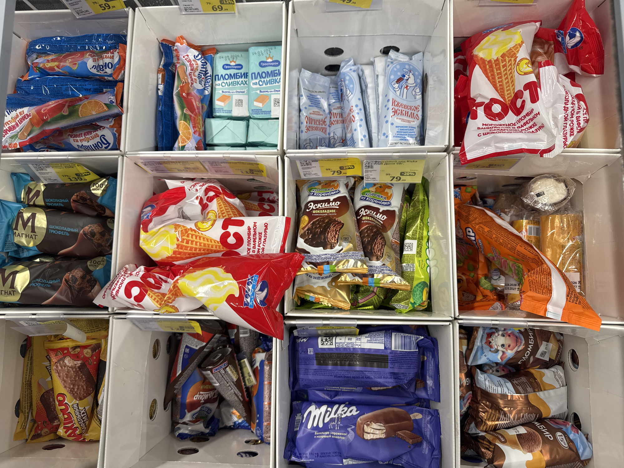 The image shows a freezer display case filled with various types of ice cream products. The display is organized into a grid of rectangular compartments. Each compartment contains several individually packaged ice cream bars, cones, or multipacks. In the top left compartment, there are several yellow and brown wrapped ice cream bars, with some packaging featuring the brand name "MAXI DUO". The compartment to its right contains chocolate-covered ice cream bars with dark brown wrappers, some with gold lettering. The next compartment to the right, in the top row, has multiple blue-wrapped ice cream bars with illustrations of fruit, such as oranges. The packaging includes Russian text. In the second row, left compartment, there are packages of Milka brand chocolate ice cream bars, identifiable by their purple packaging and the Milka logo. The compartment to the right of the Milka bars contains more ice cream products, including some with waffle cones and red and yellow packaging. The third row, left compartment, displays chocolate ice cream bars with wrappers featuring illustrations of a cow and the word "Эскимо" (Eskimo). The compartment to the right of the "Eskimo" bars contains a variety of ice cream products, many with blue and white packaging and illustrations, also featuring Russian text. Some appear to be ice cream bars. The bottom row, left compartment, contains several large, foil-wrapped ice cream products, some with images of cows and the word "ПЛОМБИР" (Plombir). The compartment to the right of the "Plombir" products contains more ice cream cones and bars, including some with red and white packaging and waffle cone designs. The far right compartment of the bottom row shows a variety of ice cream products in red and white packaging, as well as some loose ice cream. The overall environment appears to be a retail setting, likely a supermarket or convenience store freezer section, with the products neatly arranged for sale. There are price tags and labels visible on some of the products, and some of the packaging has dates and nutritional information. The image is taken from a slightly elevated angle, looking down into the display case. No people are visible in the image. The branding and text on the packaging suggest the location is likely in Russia. The question asks to identify the city of Perm, Russia, but there is no specific visual information in the image that confirms this location. However, the presence of Russian