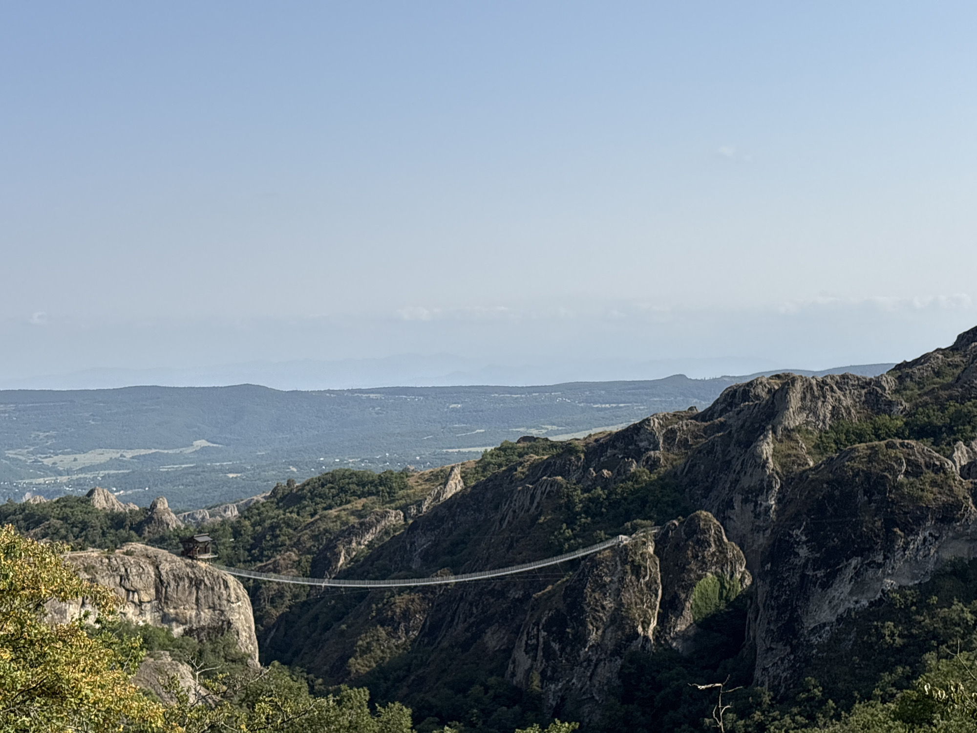 The image depicts a dramatic landscape in Orbeti, Georgia, featuring a long, slender suspension bridge spanning a deep canyon between rugged, rocky cliffs. The bridge appears to be made of metal and stretches from one sheer rock face to another. On one of the cliffs, a small, dark, wooden structure with a pitched roof is perched precariously. The surrounding terrain is heavily forested with lush green trees covering the slopes of the mountains and filling the canyon below. In the far distance, rolling hills and a hazy valley with a scattering of buildings can be seen under a clear, pale blue sky. The overall scene conveys a sense of natural grandeur and isolation.