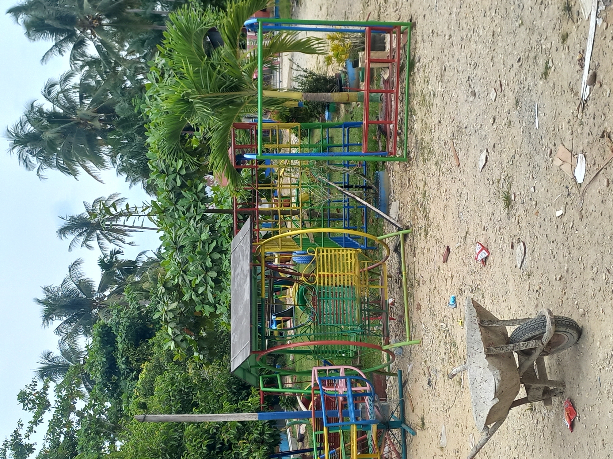 The image depicts an outdoor scene in Gampongkeude, Indonesia. The primary subjects are a colorful, multi-level playground structure made of metal tubes painted in red, green, blue, and yellow, and a weathered concrete wheelbarrow resting on a sandy ground. The playground appears to be unoccupied.
The setting is an open, sandy area with scattered debris, suggesting it might be under construction or in a less-maintained state. Lush green foliage and palm trees surround the playground, indicating a tropical environment. The sky is overcast, suggesting it might be daytime but with cloudy weather.
The playground structure features various components, including what looks like a slide, climbing frames, and possibly swings or a roundabout, all constructed from metal. The wheelbarrow, a common tool for moving materials, is dirty and shows signs of wear and tear.
There is no visible text in the image. The overall impression is one of a rustic, tropical location with a colorful but possibly unused playground and some construction materials present. The scene is static, with no discernible activity or event taking place beyond the passive presence of the objects and the natural environment.