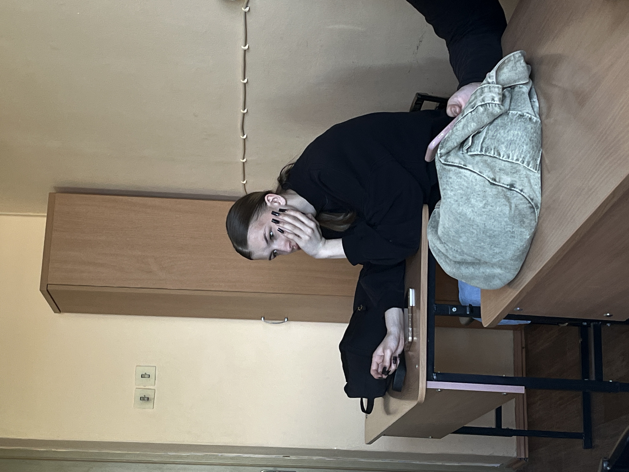 The image, rotated 90 degrees clockwise, captures an indoor scene, likely a classroom or study environment. A young woman, possibly a student, is the central subject. She has dark hair styled back and is wearing a black long-sleeved top. Her hands, with dark nail polish, are near her face, suggesting a moment of contemplation, shyness, or suppressing a smile. She is leaning on a wooden desk with a black metal frame.
Next to her on the desk, a light-colored, possibly denim, jacket is folded or draped, and a strap from a black bag or backpack is visible. In the background, a light brown wooden cabinet or shelving unit is against a light beige wall. A string of white decorative lights hangs vertically on the wall to the left of the woman. Further down on the wall, two square white electrical outlets or data ports are visible. The general setting appears quiet and informal. There is no visible text in the image.