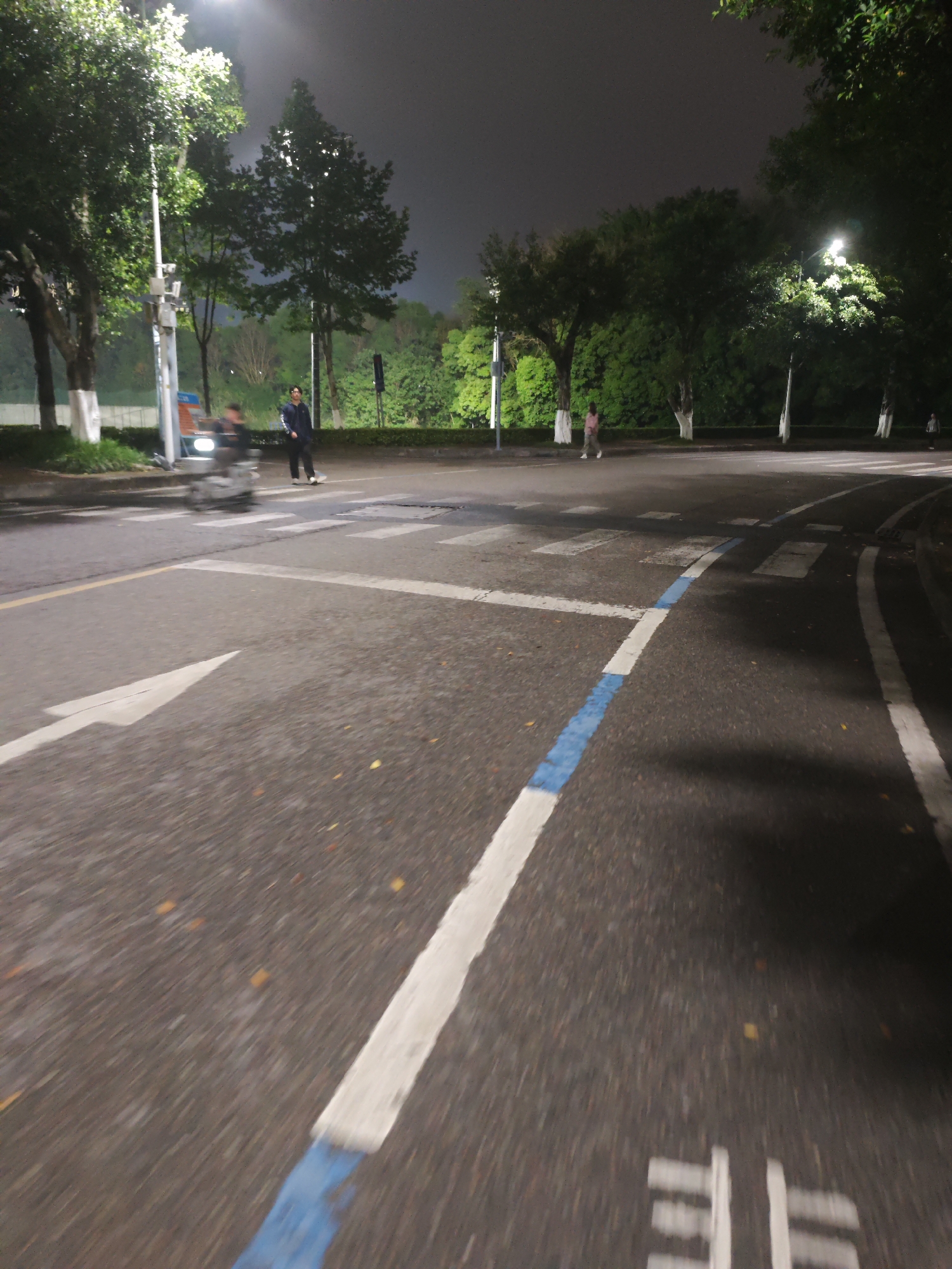 This media file depicts a night scene on a paved road in Chongqing High-tech Zone, China. The road features clear white traffic markings, including a large directional arrow in the foreground and a crosswalk further up. A distinctive blue line runs parallel to a white line, segmenting the road. Near the bottom right, a partially visible Chinese character "非" is painted on the road, likely part of "非机动车道" (non-motorized vehicle lane).
The scene is illuminated by bright streetlights, which highlight the lush green foliage of trees lining both sides of the road. The sky above is dark, indicating nighttime.
Several individuals are present. On the left, a person wearing dark clothing stands near the crosswalk. Next to them, a motor scooter or electric bike is captured in motion, appearing blurred. In the background, another person walks on the right side of the road, and a smaller, more distant figure is also visible near the trees. The overall impression is a bustling urban street at night.