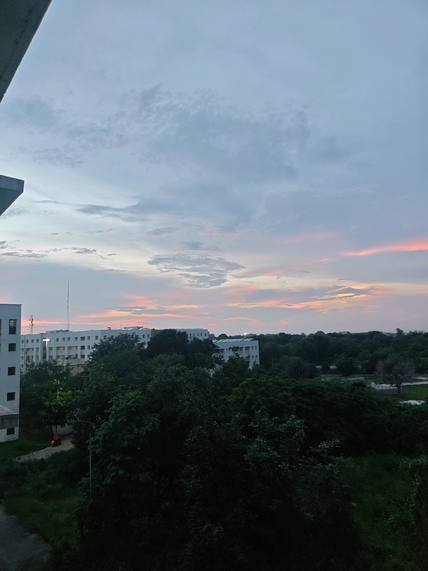 This image captures a serene evening sky over Datia, India, as twilight begins to settle. The sky is a canvas of soft blues and grays, streaked with delicate hues of pink and orange from the setting sun. Wispy clouds drift across the horizon, adding texture and depth to the celestial display.
Below the sky, the landscape is dominated by lush greenery, with dense trees forming a natural canopy. Beyond the trees, a series of modern buildings, likely apartments or educational institutions, are visible. These structures are predominantly white with dark window frames, and some feature balconies. A tall communication tower stands prominently among the buildings, its slender form reaching towards the sky. In the lower left of the image, a portion of a building's exterior, constructed with concrete and featuring dark windows, is visible. A paved pathway winds through the greenery, and a solitary red vehicle is parked on it, its red taillights faintly visible in the dimming light. The overall scene suggests a quiet, urban fringe environment where nature and infrastructure coexist.