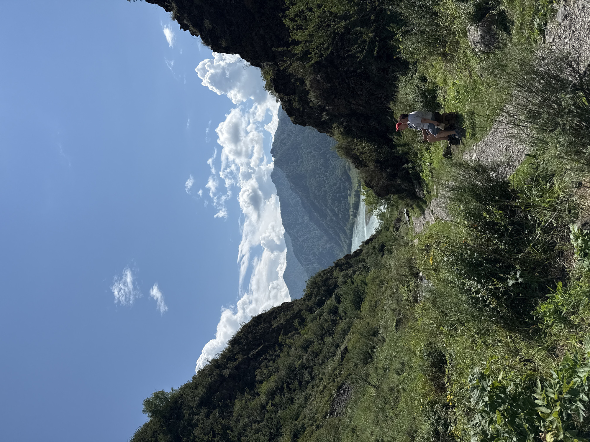 The image depicts a scenic mountain landscape with a river winding through a valley. Two individuals are seated on a log on a dirt path, overlooking the vista. Lush green vegetation covers the slopes of the surrounding mountains, with a clear blue sky dotted with white clouds overhead. The scene appears to be a tranquil outdoor setting, possibly a hiking trail or a viewpoint. Based on the overall aesthetic and the potential for such landscapes, this scene is likely located in the vicinity of Chemal, Russia, a region known for its natural beauty and mountainous terrain.
