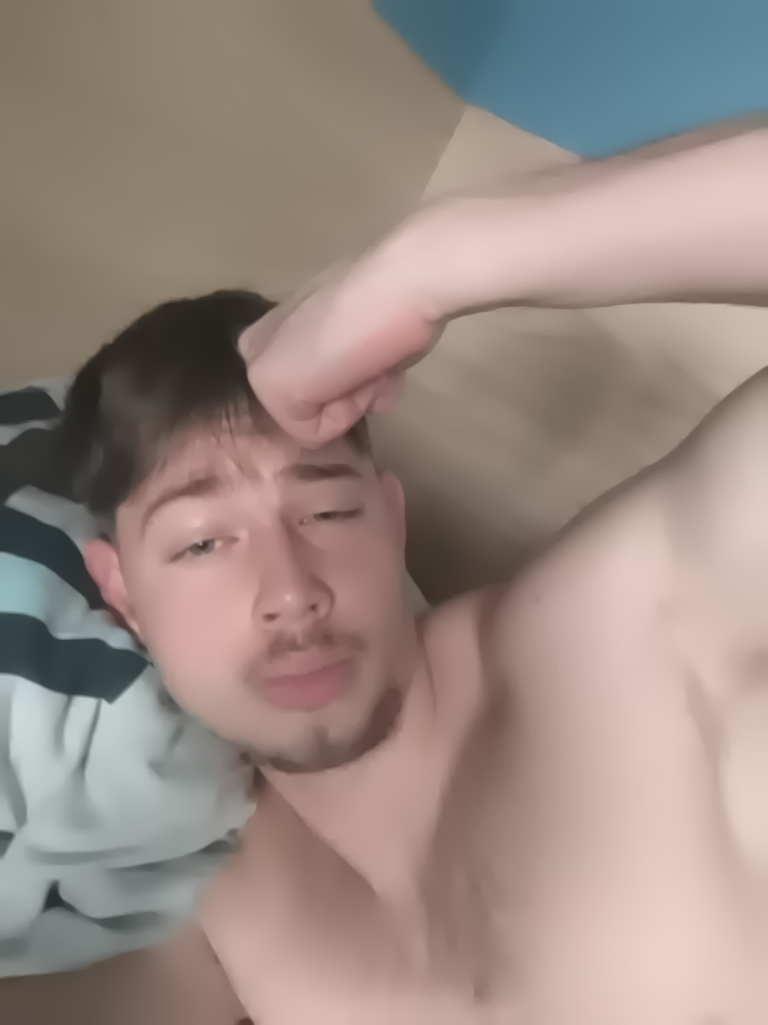 A Caucasian male with dark hair and a trimmed beard lies on a pillow with green and white stripes. His torso is bare, and he rests his fist on his forehead. The background shows a beige wall transitioning to a blue wall in the upper right corner. This image does not contain information about a location in Sofia, Bulgaria.