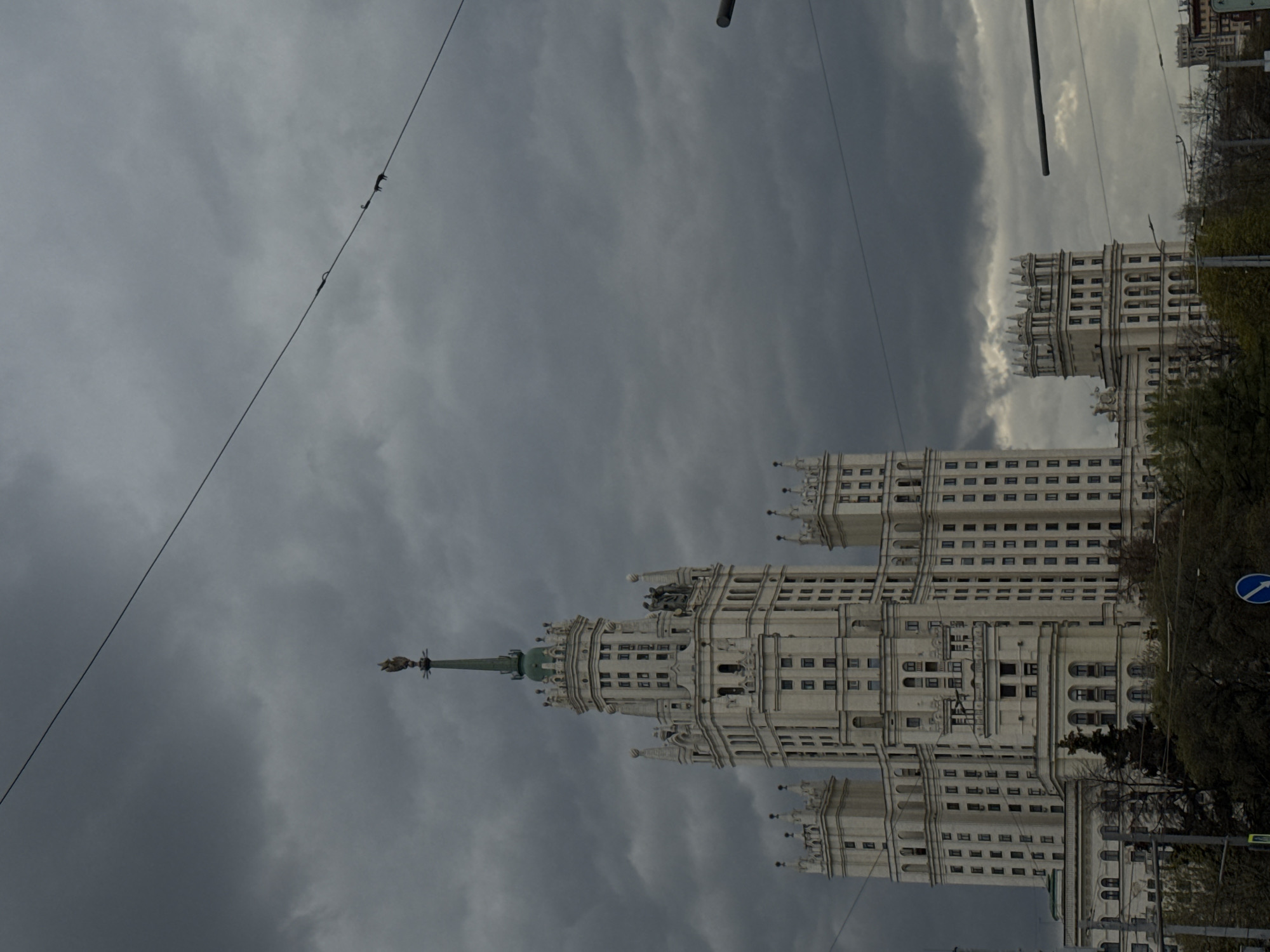The image captures a grand, ornate Stalinist skyscraper, characteristic of Moscow's "Seven Sisters," under a heavily overcast sky. The monumental building, likely the Kotelnicheskaya Embankment Building, is seen from a low angle, emphasizing its height and intricate architecture. It features multiple tiers, countless windows, and decorative elements such as turrets, cornices, and statuary. A prominent green spire, topped with a golden finial (possibly a star), crowns the central section of the structure.
The sky is filled with dark, dramatic grey clouds, suggesting a somber day, possibly impending rain or heavy cloud cover throughout the day. Power lines stretch across the upper and left portions of the frame, with several insulators or connectors visible on them. To the right and at the bottom of the building, patches of green trees and foliage are visible. A blue road sign with a white arrow pointing to the right, indicating a "turn right ahead" or "one way" instruction, is partially visible on the right side. The scene is devoid of people or specific activity, focusing solely on the architectural landmark and the dramatic weather conditions in Moscow, Russia.