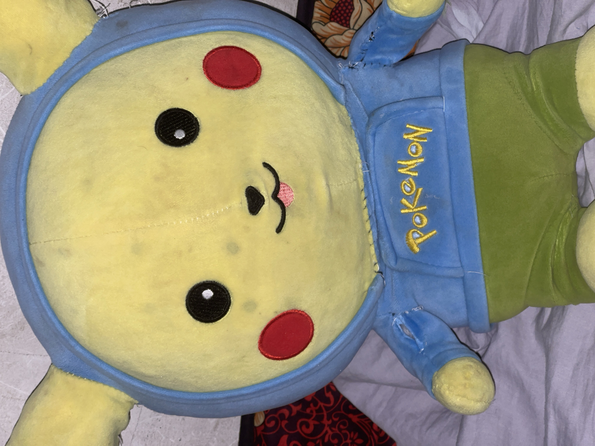 The image shows a close-up of a plush toy resembling Pikachu, a character from Pokémon. The toy is yellow with black eyes, red circles on its cheeks, and a small pink tongue sticking out. It is wearing a blue hoodie with "Pokemon" embroidered in yellow text on the front and green shorts. The toy is positioned on a bed with a light purple blanket. In the background, a wall with a textured surface and a glimpse of a patterned fabric are visible. The location is specified as a domestic setting in the city of Кызыл-Тоо, Kyrgyzstan. There are no people or actions depicted in the image.