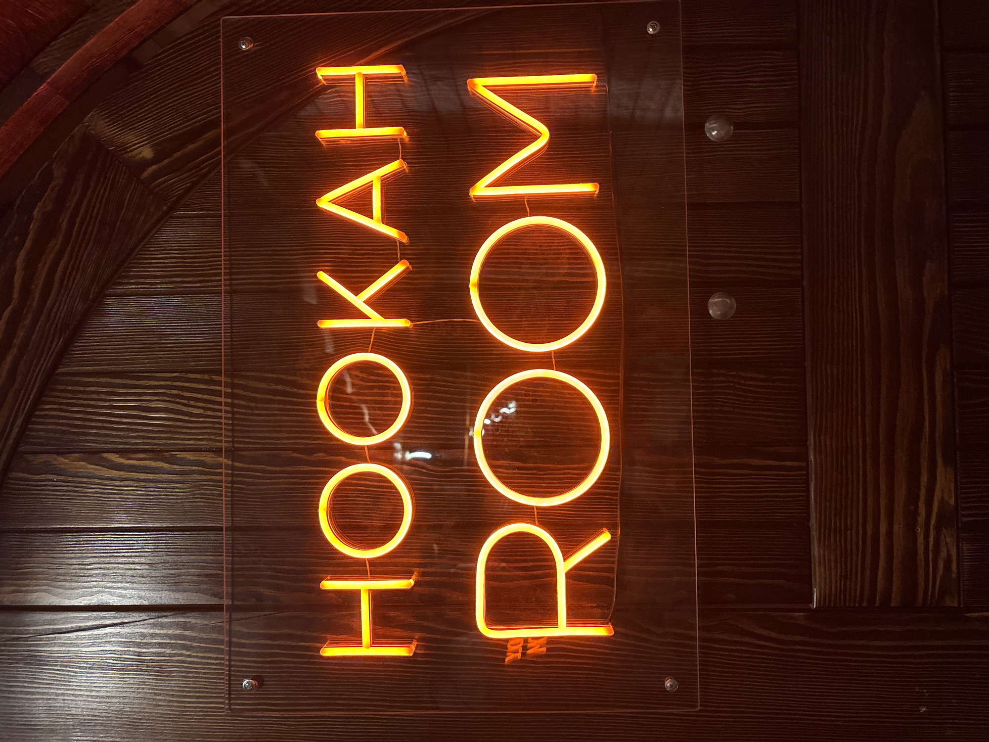 The image shows a neon sign with orange lettering that reads "HOOKAH ROOM" vertically. The sign is mounted on a dark wooden wall, which appears to be made of horizontal planks. The neon lights cast a warm glow on the wood. The sign is enclosed in a clear acrylic sheet, secured to the wall with metal screws. The lighting suggests an indoor setting, possibly a lounge or bar, and the time of day is not discernible. The overall impression is one of an inviting, dimly lit establishment. The scene is static, with no people or activity depicted. The focus is solely on the sign and its immediate surroundings. The location is identified as Chaikovsky, Russia.