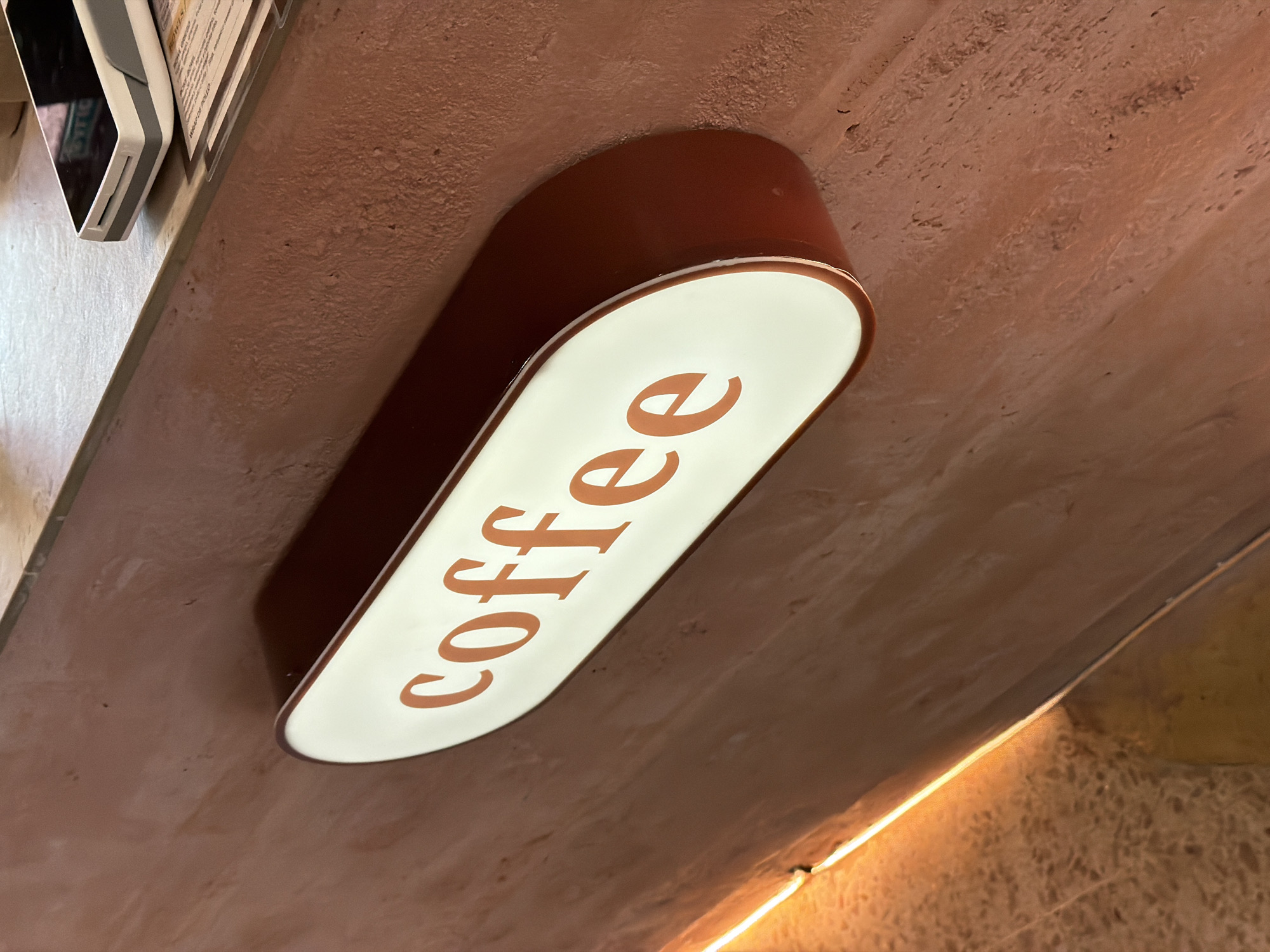 An illuminated sign with the word "coffee" in orange font on a white background is affixed to a textured reddish-brown wall. The sign has a dark red border and casts a shadow on the wall. Below the sign, a strip of warm-toned lighting illuminates the bottom portion of the wall and a speckled floor. In the upper right corner, part of a credit card reader and some papers are visible on a counter. The setting appears to be a cafe or coffee shop. This scene is located in Benidorm, Spain.