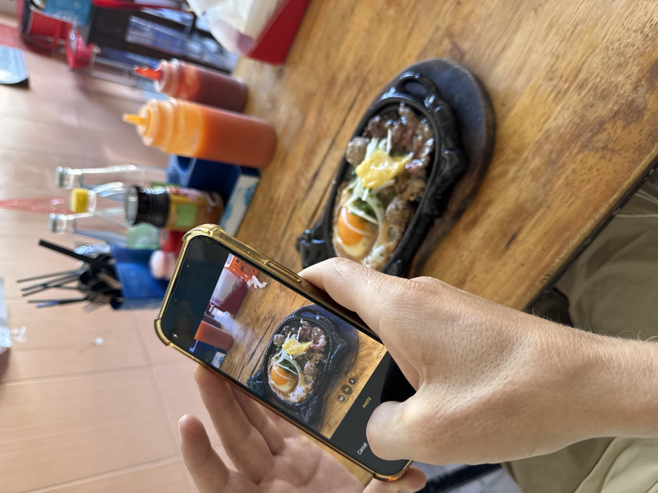 A person holds a gold-cased iPhone, displaying a picture of food in a black, oval serving dish. The food appears to include eggs, meat, and shredded toppings. The iPhone's camera interface is visible. The image is being captured on a wooden table alongside condiments like red and orange sauces in plastic bottles. Utensils are placed in a blue container in the background. Part of the person's arm and shirt are visible on the right. The floor has a light-colored wood pattern.