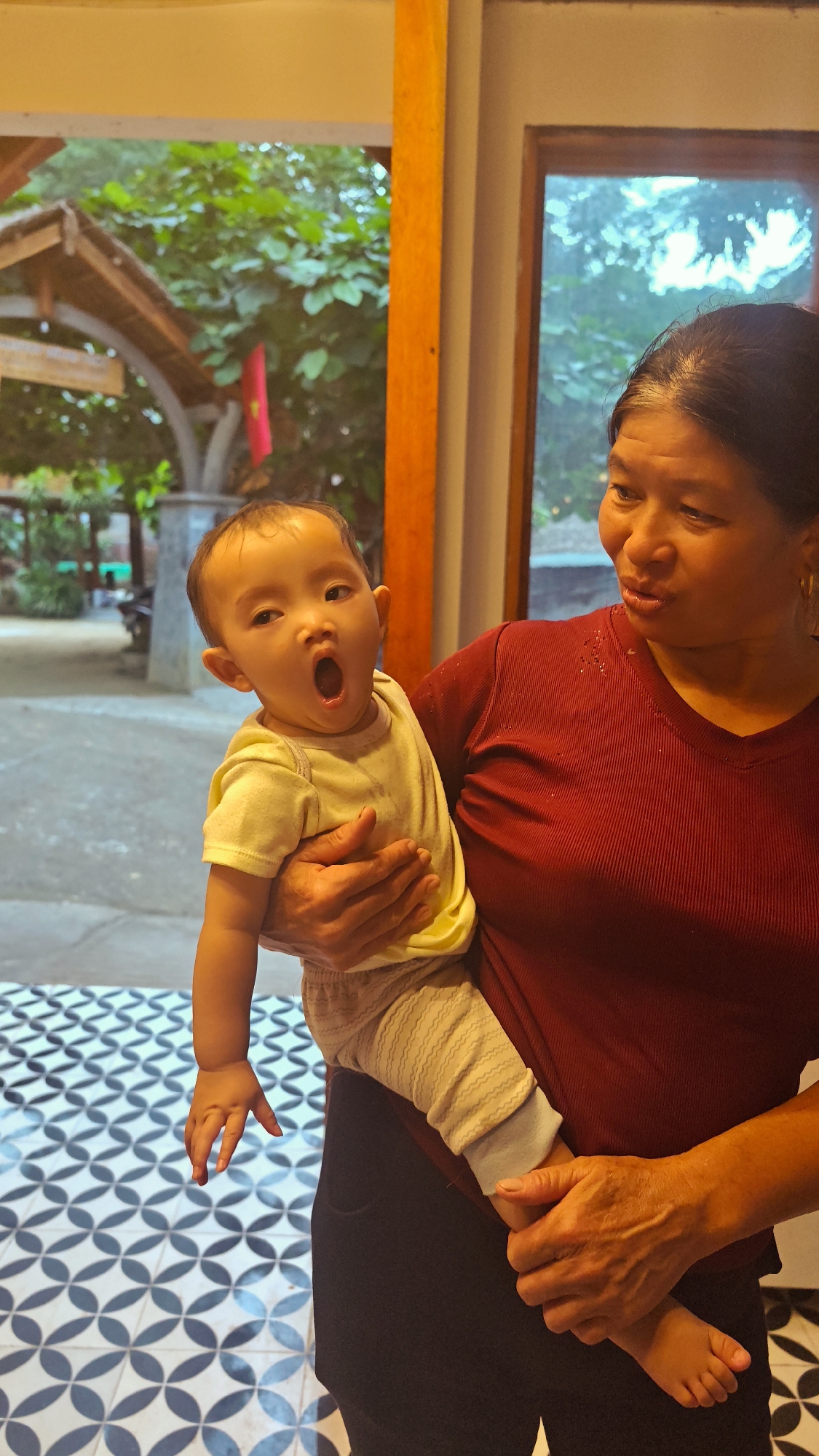 An East Asian woman in a dark red, ribbed V-neck shirt holds a baby in Cổ Lũng Commune, Vietnam. The baby, wearing a light yellow shirt and grey patterned pants, is held with their mouth open in a yawn and their eyes looking towards the camera. The woman is looking slightly to her right, with her head tilted. Her hands cradle the baby. Behind them, a large window reveals an outdoor scene with lush green trees and a structure with a thatched roof. The foreground features a black and white patterned floor.