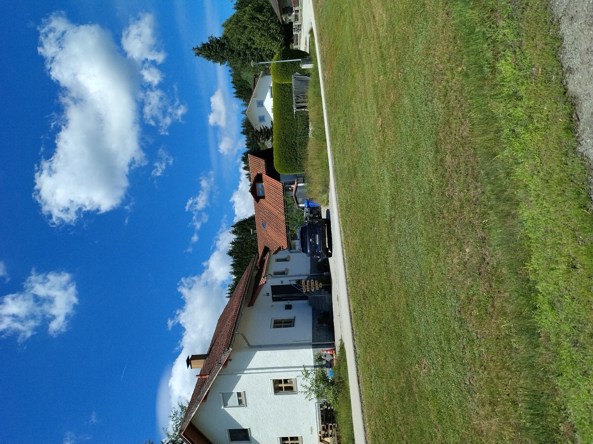 The image depicts a residential scene in Freyung, Germany, under a bright blue sky with scattered white clouds. In the foreground, a grassy lawn stretches across the frame, with a gravel path or road bordering the bottom right. Several houses are visible in the background, characterized by red tiled roofs and white or light-colored exteriors.
The most prominent house on the left is a two-story white building with several windows and a chimney. A dark blue car is parked on the driveway in front of this house. Adjacent to the car, a multi-directional wooden signpost is erected, indicating various destinations such as "Zuhause" (Home), "Maria," and other unreadable names. To the right of this house, another residence with a red tiled roof and dormer windows is partially visible, with a manicured green hedge in front. Beyond these houses, a dense forest of evergreen trees forms the backdrop.
The environment appears to be a suburban or rural area, with well-maintained properties and natural landscaping. There are no people clearly visible engaging in any specific actions. The overall impression is one of a peaceful, sunny day in a German countryside setting.
