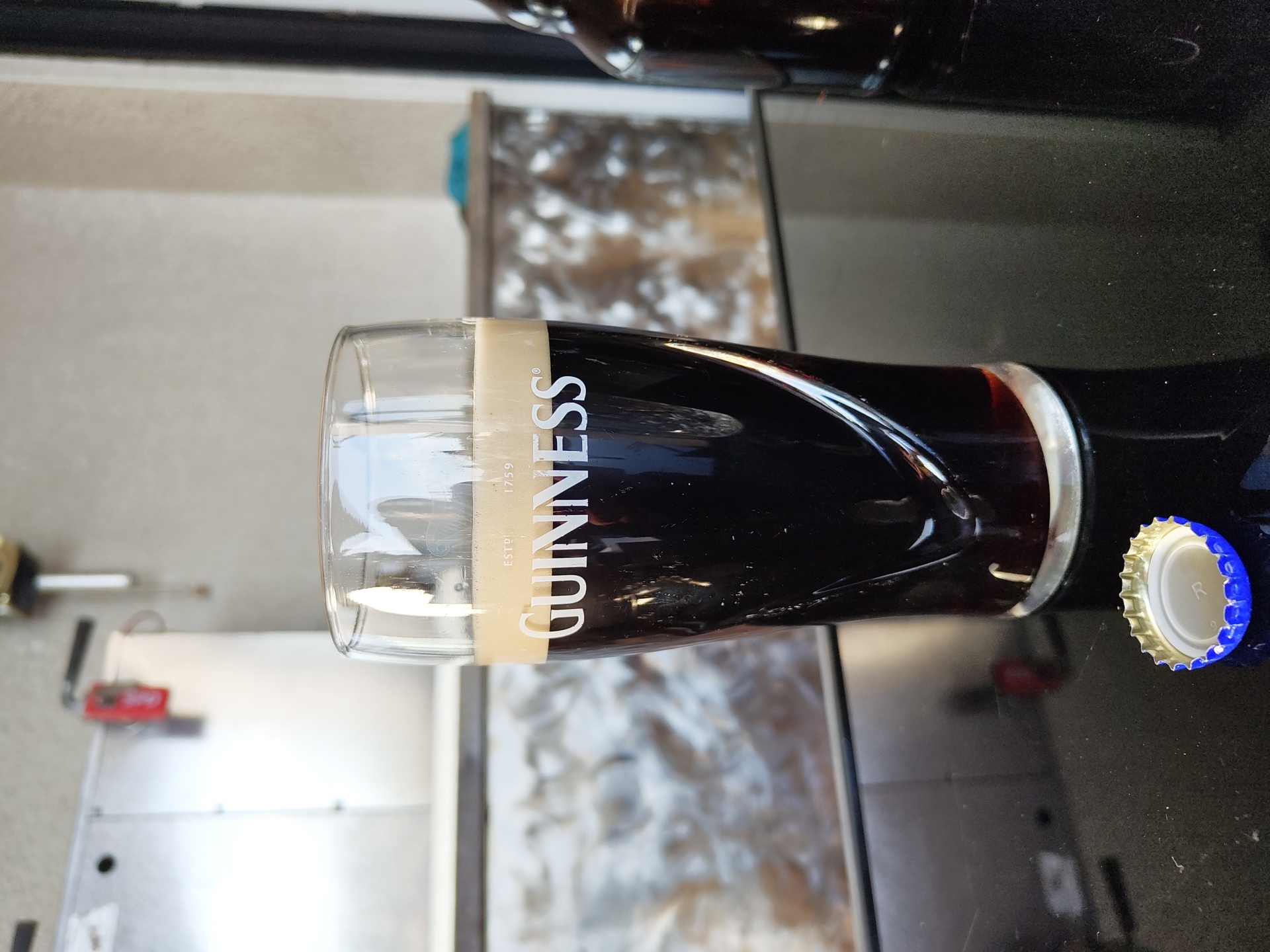 The image depicts a filled Guinness glass with a bottle of Guinness next to it. The background includes a countertop and an out-of-focus area showing a possible service area. The location cannot be determined to be Schmelz, Germany with the information provided.