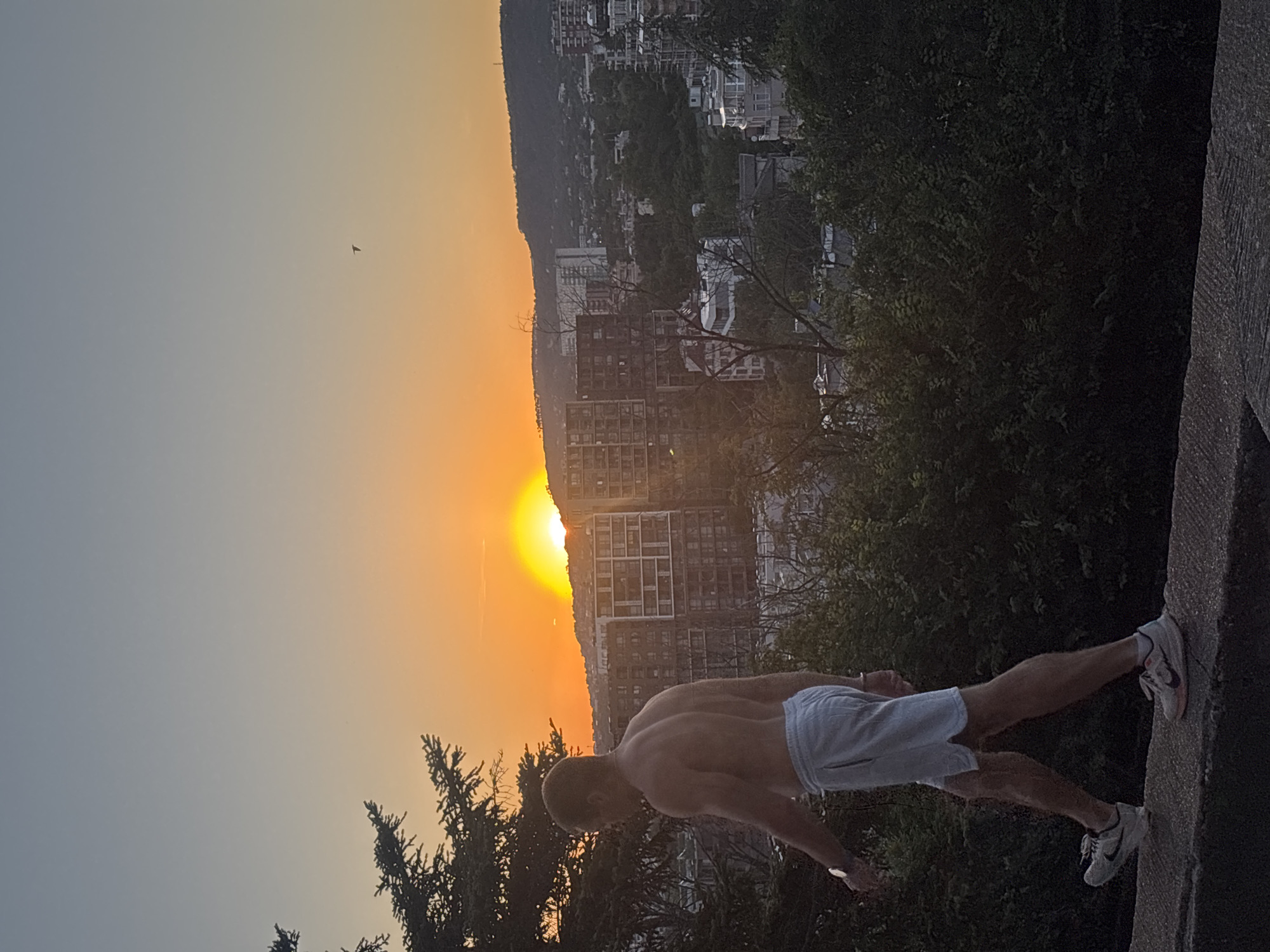 In Varna, Bulgaria, as the sun sets, a man walks along a stone ledge, his back to the camera. He is shirtless, wearing light-colored athletic shorts and white sneakers. The golden light of the setting sun illuminates the sky and the urban landscape behind him. The city is characterized by multi-story buildings and lush greenery, with a distant hill forming the backdrop. The man's posture suggests a leisurely walk or a moment of contemplation as he observes the sunset. The scene is peaceful, capturing the transition from day to evening in a coastal city.