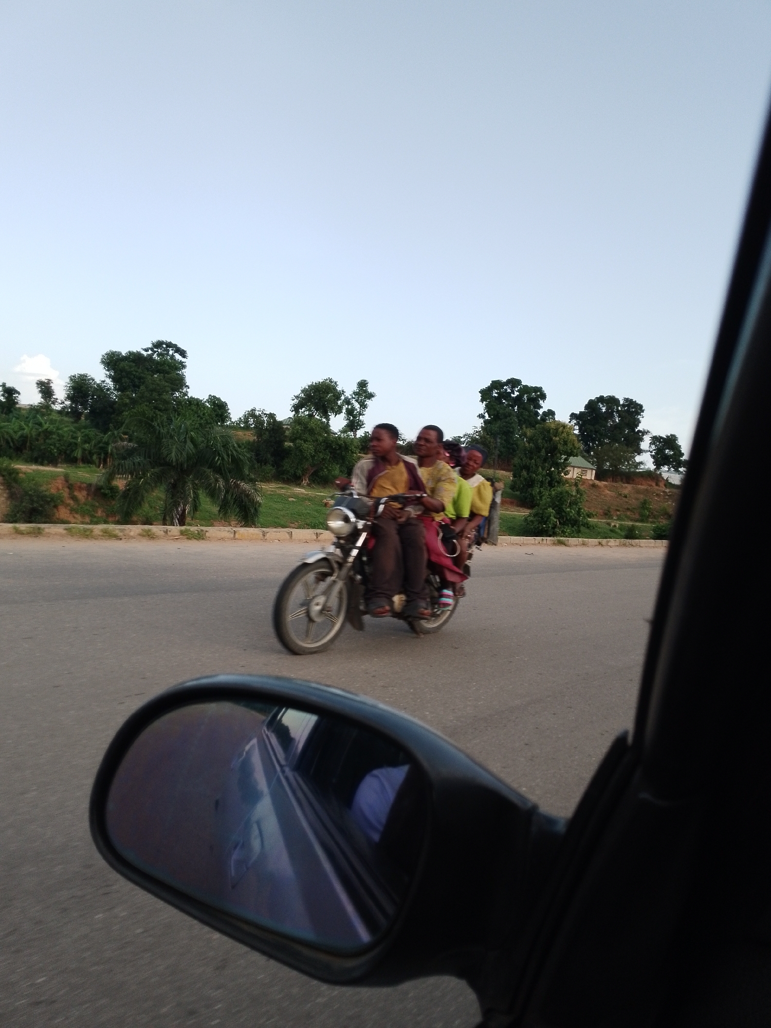The image shows a motorcycle with four people riding on it, traveling on a paved road. A person in a light-colored shirt is driving, while three passengers, two adults and a child, are seated behind them. The motorcycle is silver and red. The background consists of a grassy embankment with trees and a house. The scene appears to be in a rural or semi-rural area. The photograph is taken from a moving vehicle, as evidenced by the side mirror in the foreground reflecting the road and sky.