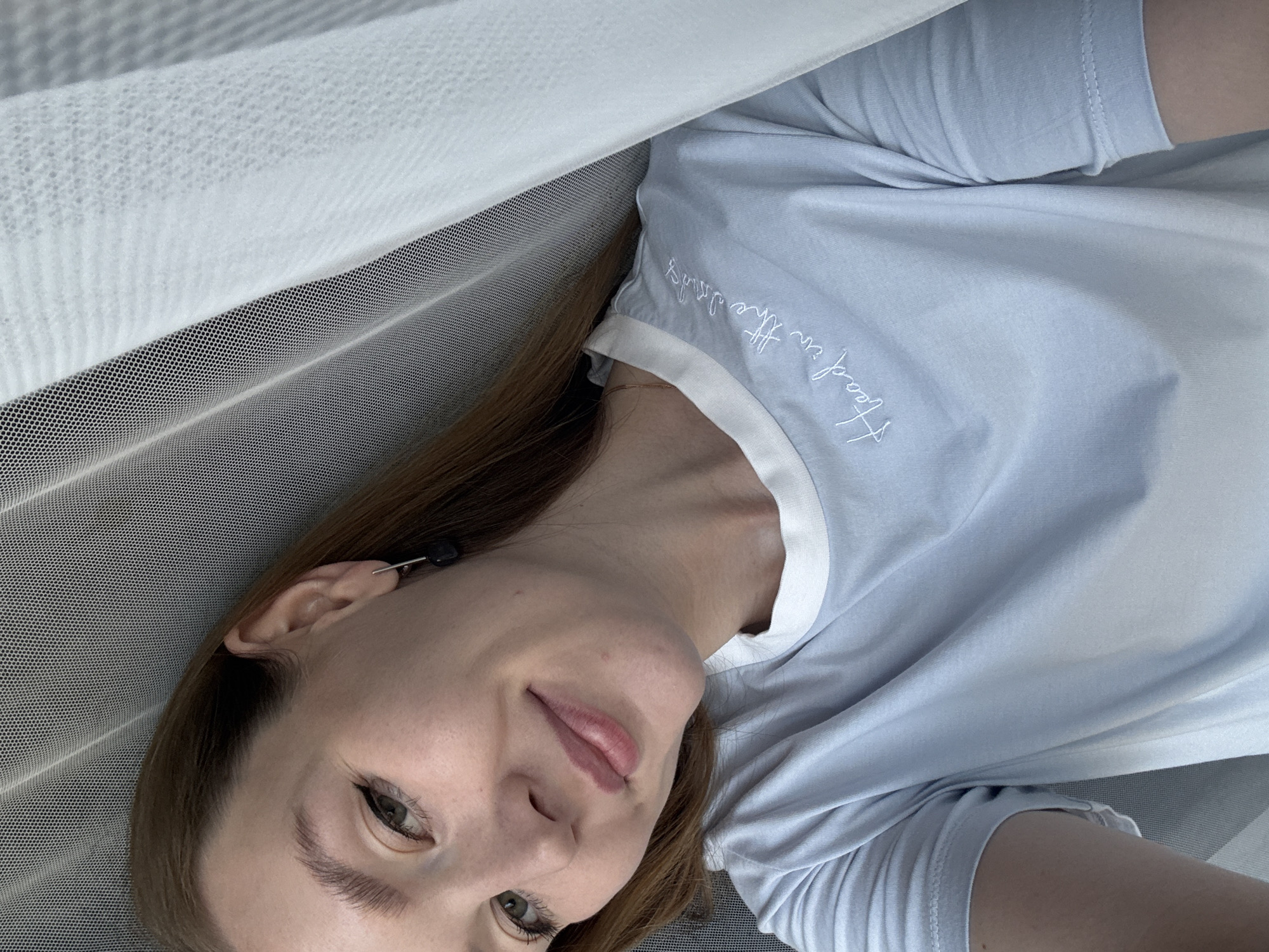 A woman with shoulder-length brown hair and green eyes, wearing a light blue t-shirt with white trim and embroidered text that reads "Head in the clouds," is pictured indoors. She is leaning her head against a sheer white fabric, possibly a curtain or netting, with a gray surface visible behind her. Her makeup includes eyeliner and lip gloss. She is wearing a dark earring in her right ear. The scene is a close-up shot of her face and upper torso, taken from a slightly high angle. The environment appears to be domestic. There is no identifiable information about the location within Izhevsk, Russia.