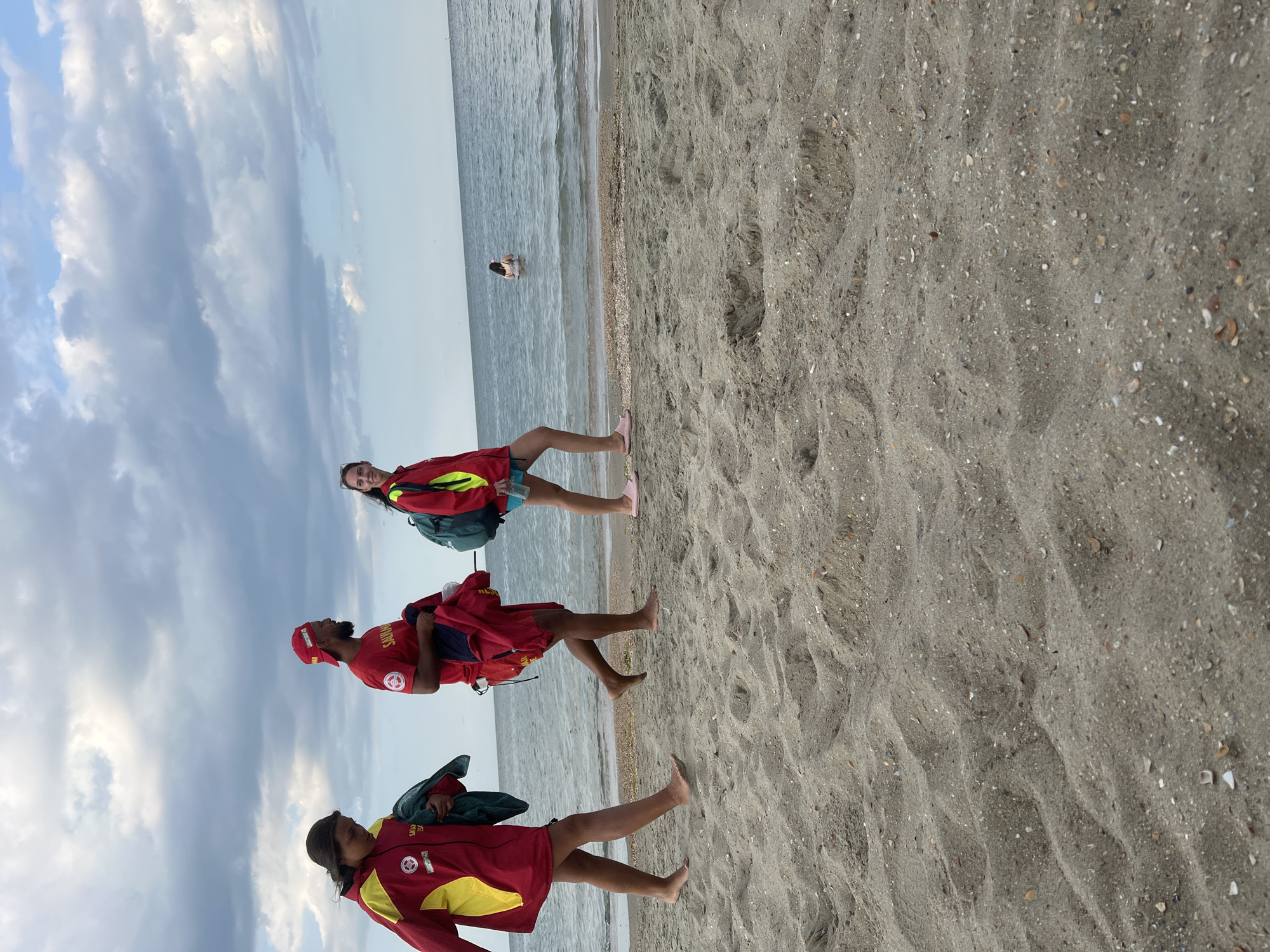 The image shows three people on a sandy beach, with the sea in the background. All three individuals are wearing red jackets with yellow accents and visible text on them. One person is facing the camera, while the other two are looking away. They appear to be lifeguards, possibly on duty.
The person closest to the camera on the bottom left is wearing a red jacket and has a towel over their shoulder. The person in the middle is wearing a red jacket and a red cap with visible text. The person at the top right is also wearing a red jacket and is facing the camera.
The sea is calm with gentle waves. In the distance, a person can be seen standing in the water. The sky is partly cloudy with a mix of blue and white clouds, suggesting it might be daytime, possibly late afternoon or early morning due to the lighting. The sand is light brown and textured with small shells and pebbles.
The text visible on the jackets reads "SAVAS" and "SAVA". The scene appears to be a public beach, likely in Constanța, Romania, given the context. There are no strong indicators of emotions, but the general atmosphere appears relaxed and possibly professional given the attire of the subjects.