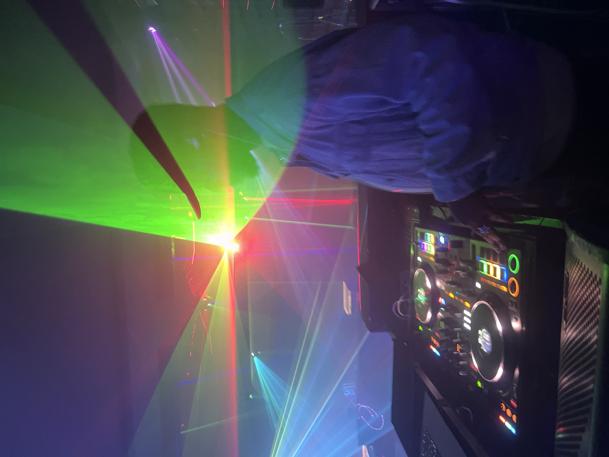 The image shows a person wearing a blue shirt standing next to a DJ mixing board at a club or event, with multiple laser lights creating a complex display of colors, including green, red, purple, and blue. The location is Unknown, Unknown.