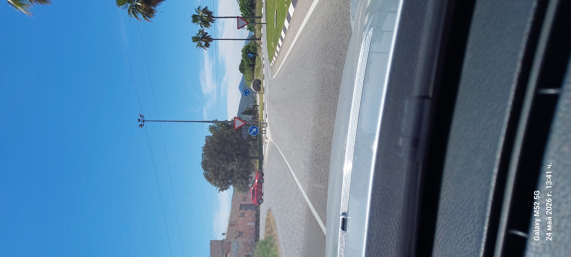 The image, taken from inside a vehicle, shows a view of a road and its surroundings in Canals, Spain. The road has white lines marking the edges and a median with vegetation. Road signs are visible, including a triangular warning sign and a blue circular sign indicating a mandatory direction of travel. Palm trees line the road. In the distance, a mountain is partially obscured by clouds. A building is visible in the background. The sky is blue. The vehicle's door is prominent in the foreground, partially obstructing the view. A red car is parked beside the road. A lamppost stands tall on the left of the road. The image was taken on May 24, 2026, at 13:41, according to the timestamp from a Galaxy M52 5G device.