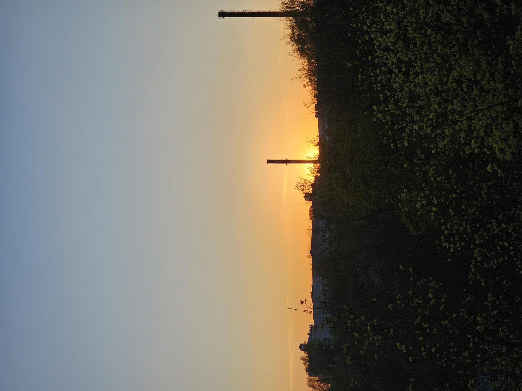 The image shows a sunset in what is most likely Khimki, Russia, suggested by the presence of two industrial chimneys. The sky transitions from a pale blue to a yellow-orange gradient at the horizon, where the sun is setting. The silhouettes of buildings and the dark green foliage of trees are visible on the right side of the image. The chimneys are vertical structures. The composition suggests the photograph was taken near or within a residential area with mature trees, overlooking a landscape with industrial elements. The image is vertically oriented.