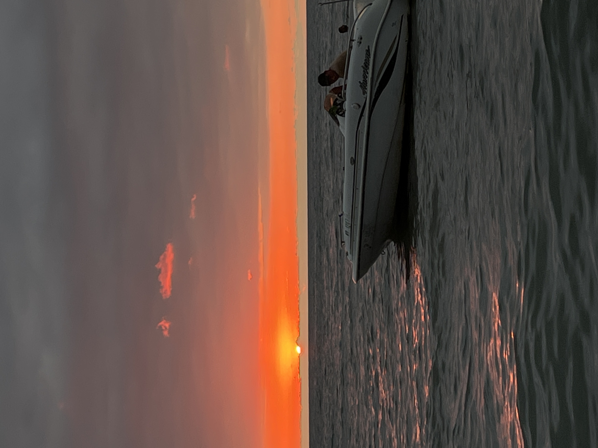 A low-angle, medium shot captures a sunset over the Black Sea in Ureki, Georgia. The sky is a gradient of dark gray clouds at the top, transitioning to vibrant orange and red hues near the horizon, where the sun is partially obscured by a thin cloud layer. Two small, bright orange clouds are visible in the upper left portion of the sky. The water is dark and choppy, reflecting the colors of the sunset. A white speedboat with black accents is positioned in the right half of the frame, moving away from the viewer. A person wearing a dark baseball cap and a tan shirt is seated in the driver's seat of the boat, looking towards the right. The boat's hull displays the "Hurricane" logo. The scene is serene and depicts a moment of leisure on the water during sunset.