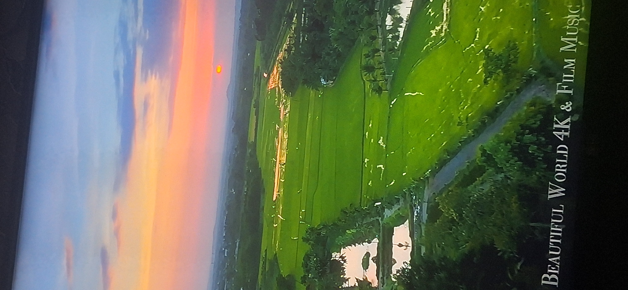 This is an aerial photograph of a rural landscape at sunset. The sky is painted with warm hues of orange, pink, and purple, with scattered clouds. Below, vast green rice paddies stretch across the land, reflecting the sky's colors. A body of water, possibly a pond or lake, is visible on the left, bordered by trees. In the distance, rolling hills and a small town can be seen. A text overlay at the bottom reads "BEAUTIFUL WORLD 4K & FILM MUSIC". The scene depicts a serene and picturesque natural setting. There are no people or specific urban features visible that would indicate a location within Bila Tserkva, Ukraine.