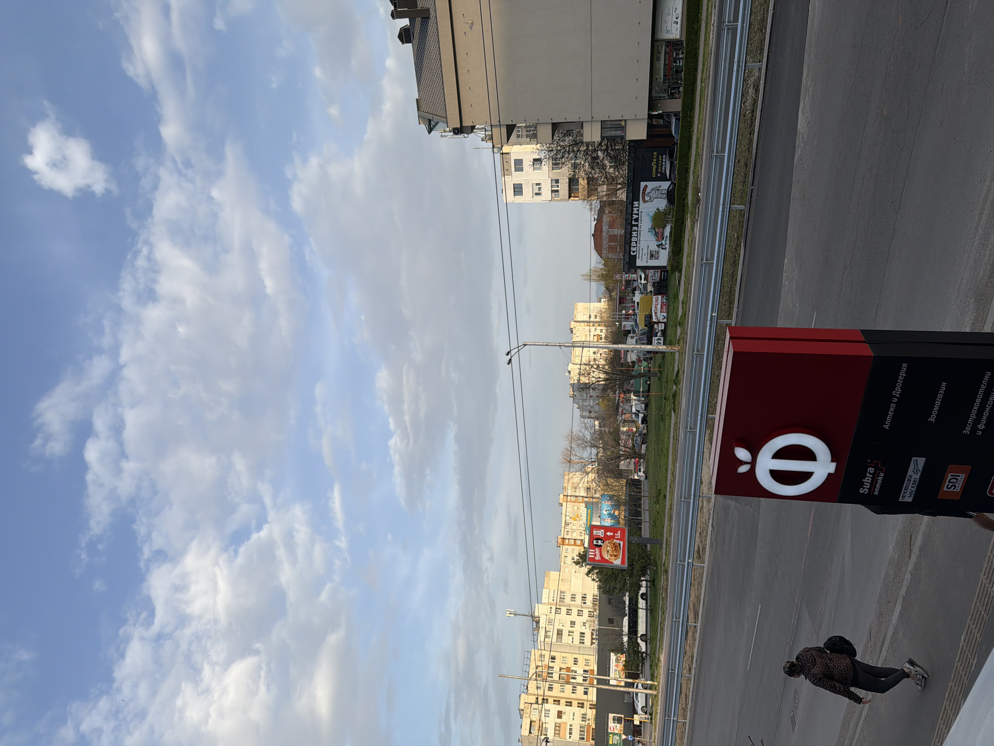 The image shows a street scene in Sofia, Bulgaria. The street is lined with buildings of varying heights and architectural styles, and there is a sidewalk and a road with vehicles. A pedestrian is walking on the sidewalk near a sign that reads "Subra" and also has the words "Аптека и Дрогерия" (Pharmacy and drugstore) on it. The sky is partly cloudy. A sign advertising "СЕРВИЗ ГУМИ" (Tire Service) is visible in the background, also advertising Goodyear, Dunlop, and Falken tires.