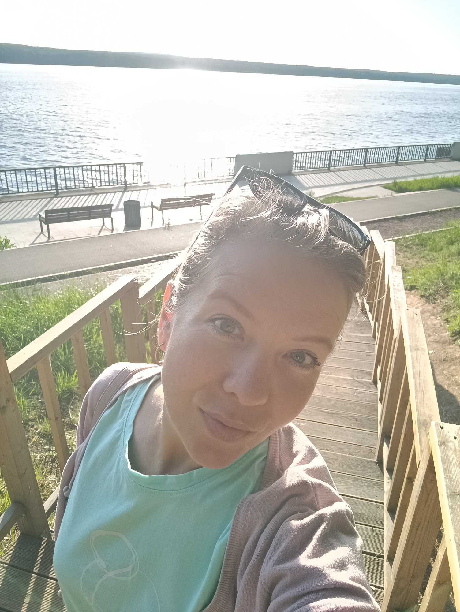 A woman is taking a selfie on a wooden staircase. She is wearing sunglasses on her head, a light green t-shirt, and a pink sweater. Behind her is a view of the Kama Reservoir in Chaikovsky, Russia. A walkway with benches and a railing can be seen along the reservoir's edge. The weather appears to be sunny.