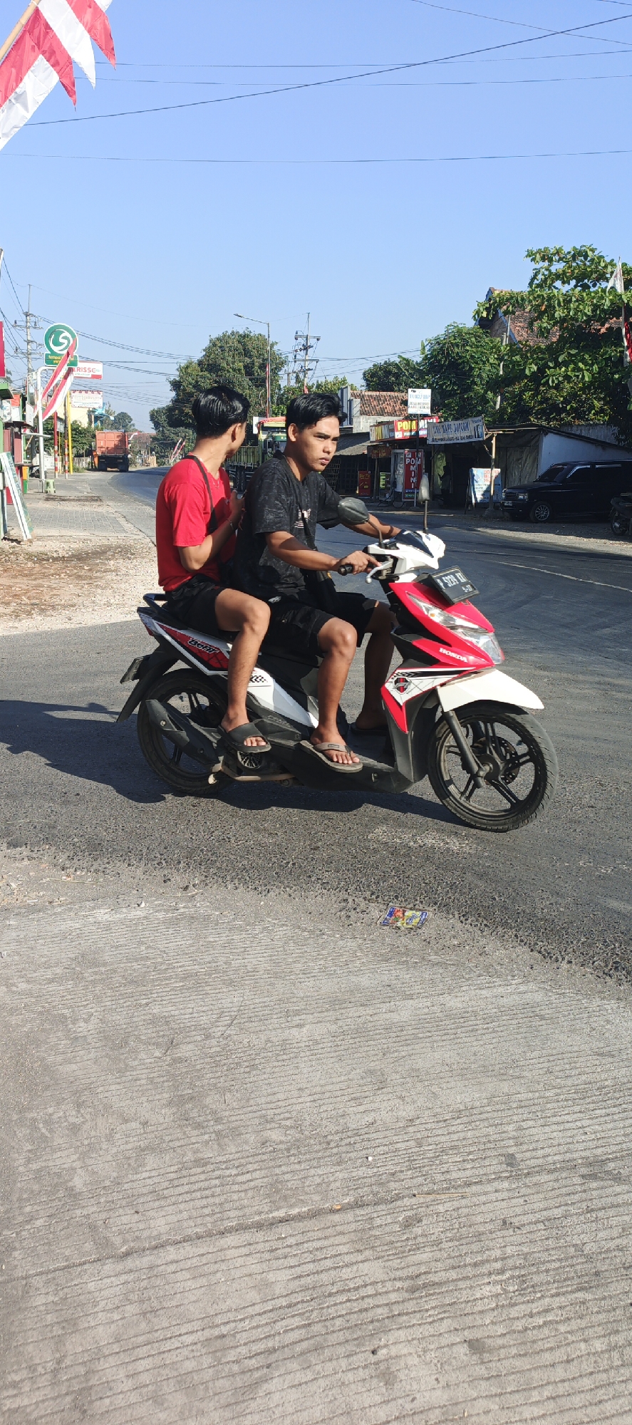 Two young males are riding on a red and white scooter on a paved road. The scooter is in motion. The driver, wearing a black t-shirt and shorts, is holding the handlebars and looking forward. The passenger, wearing a red t-shirt and shorts, is sitting behind the driver and looking to his left. They are on a street with buildings and trees on the right side and a sidewalk with various signs on the left. In the background, there is a truck and more buildings. The sky is clear and blue. The image was taken in Kasiyan, Indonesia.