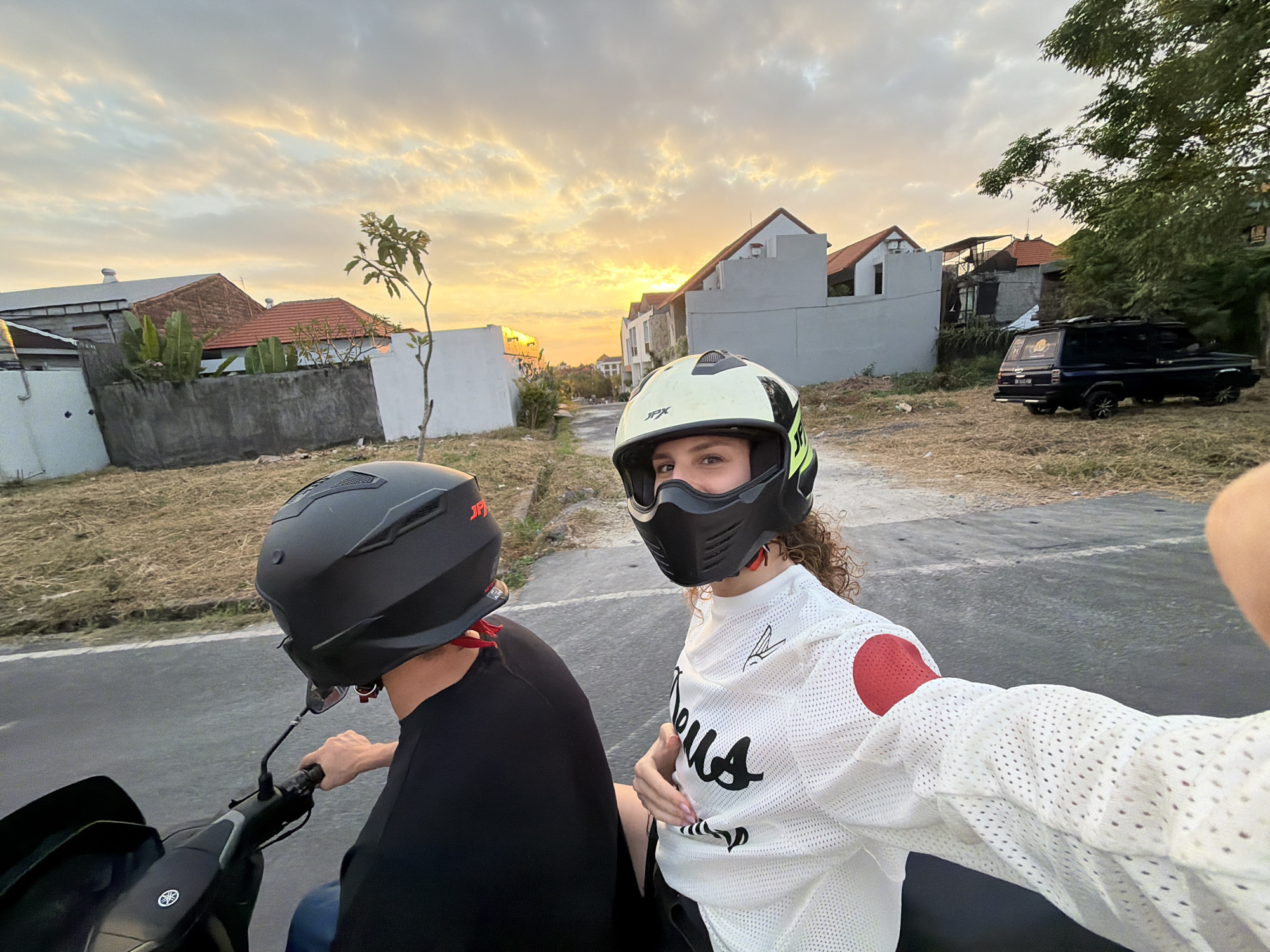 In North Kuta, Indonesia, two individuals ride a motorcycle during sunset. The person in the foreground, wearing a white mesh long-sleeved shirt with red circular accents on the shoulders and a black and green helmet, is taking a selfie. The second person, in the background and in the rider's position, wears a black helmet and a black t-shirt. They are on a paved road with dry grass and bushes on the side, bordered by residential buildings and a dark-colored SUV parked in a lot. The sky is illuminated by the warm glow of the setting sun, with scattered clouds.