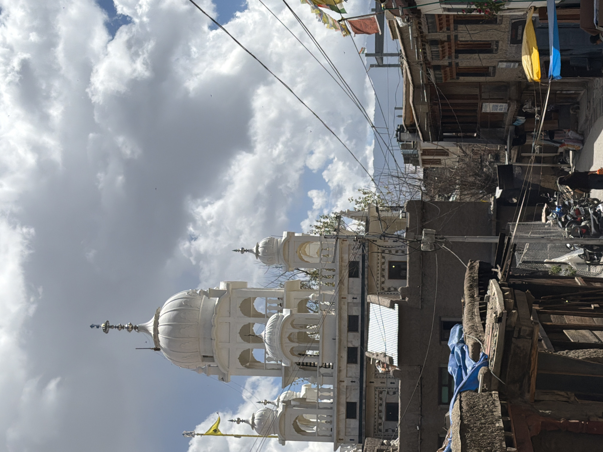This photograph captures a scene in Leh, India, featuring a prominent white Gurudwara with multiple domes and ornate architectural details. A yellow flag with a symbol is visible on a flagpole to the left of the Gurudwara. The Gurudwara is situated above a series of rustic, multi-story buildings constructed from earth and wood, with corrugated metal roofs and balconies. In the foreground, a narrow, unpaved street is lined with these traditional structures. Several motorcycles are parked on the street, and a few individuals are present, including a woman walking away from the camera carrying an orange bag. Wires and cables crisscross the sky above the street. In the background, a mountain range is faintly visible under a cloudy sky. Prayer flags, predominantly in red and yellow, are strung across some of the buildings. The overall impression is of a street scene in an older part of Leh, with a significant religious building overlooking the residential and commercial structures.