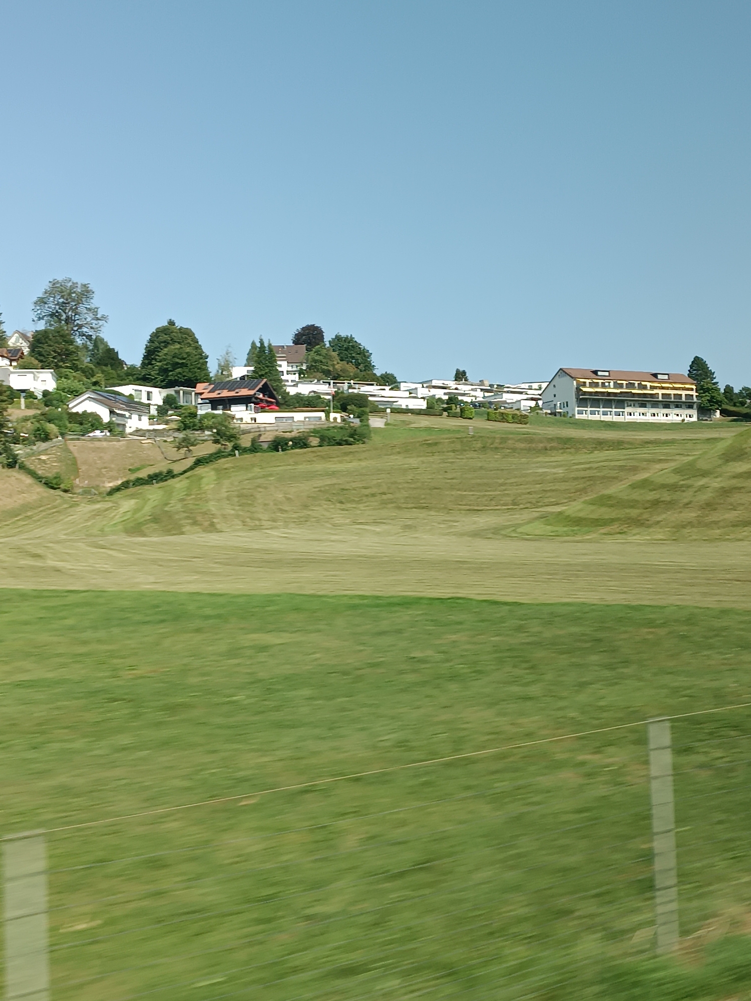 The image shows a hillside landscape with buildings and green fields. In the distance, on the crest of the hill, are several buildings, including what appears to be a hotel or large residential complex with a brown roof and multiple stories. There are also smaller houses scattered among trees. The hillside is covered in grass, with some areas mown and others longer. The sky is clear and blue. The foreground shows a blurred view of a grassy field with a fence. The location is identified as Niederteufen, Switzerland.