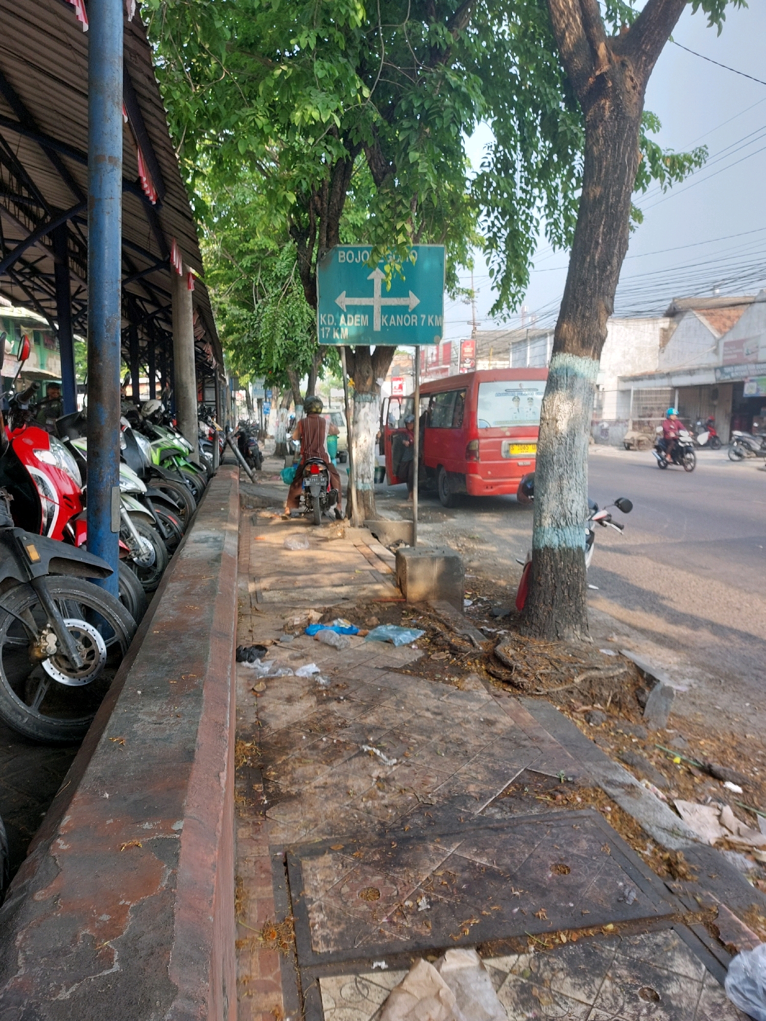 The image captures a street scene in Sumuragung, Indonesia, on a clear day. In the foreground, a row of parked motorcycles is visible, with a particularly striking red and black one in the left portion of the frame. The motorcycles are parked under a covered structure, supported by blue and grey poles, suggesting a marketplace or parking area. A person is seated on a motorcycle further down the row. A sidewalk runs alongside the parked motorcycles, marked by a concrete barrier and littered with debris including plastic bags, leaves, and other refuse. A man on a motorcycle is seen on the sidewalk, heading away from the viewer. A red van is parked on the street to the right of the sidewalk. A green directional signpost is mounted on a pole, indicating directions to "BOJO," "KD. ADEM," and "KANOR." Trees with dense green foliage line the street and sidewalk, with exposed roots visible near the base of a large tree with blue and white markings on its trunk. Further down the street, other buildings and a few more motorcycles are visible. The overall impression is a busy, somewhat unkempt urban environment.