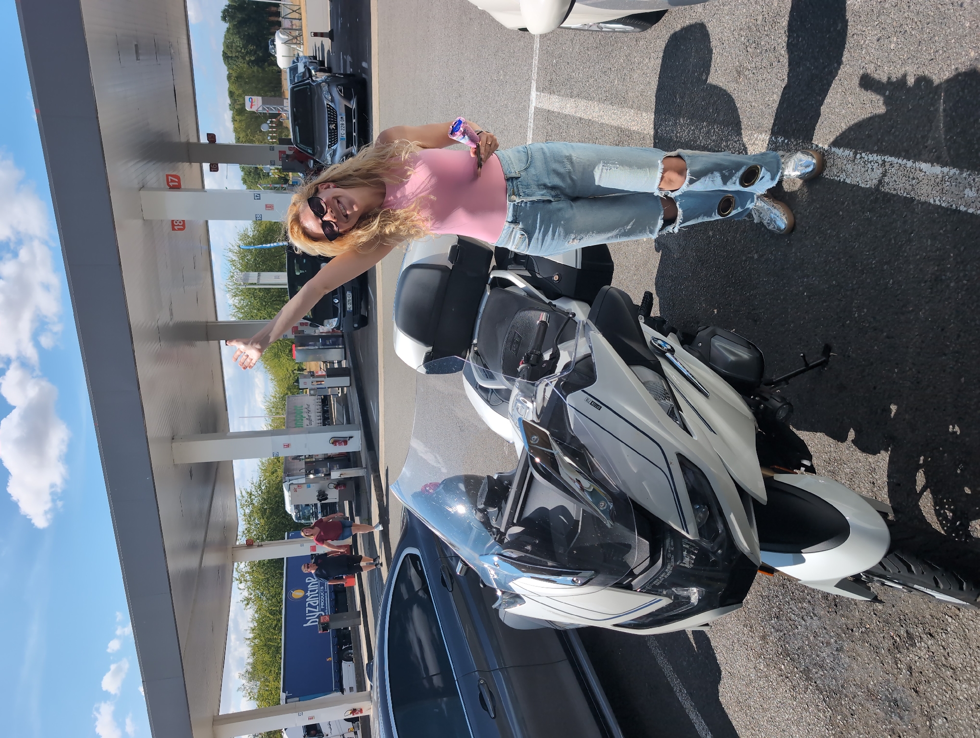 In Urvillers, France, at a gas station with a covered canopy, a woman with blonde, curly hair stands smiling and waving. She is wearing a pink tank top, distressed denim jeans with ripped knees, and silver shoes. She holds a blue and purple item in her right hand and a phone in her left. In front of her is a white and black motorcycle. Behind her, several cars are parked, and two men walk in the background near a blue truck. The sky is blue with scattered clouds.