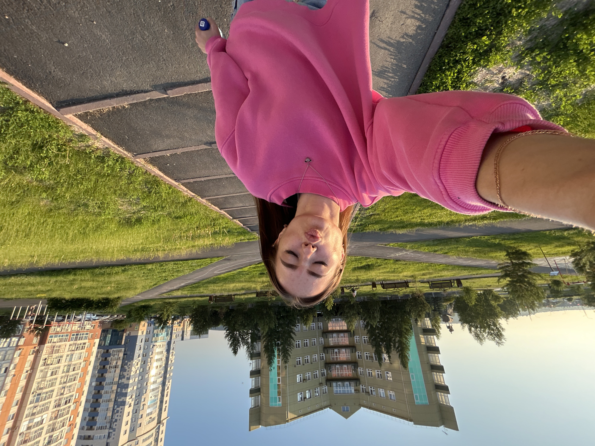 In an outdoor urban park setting during daylight, a young woman with long brown hair stands on a paved walkway, holding a phone to take a selfie. She is wearing a bright pink oversized sweatshirt and appears to be puckering her lips. The walkway is bordered by green grass and trees. In the background, two modern apartment buildings are visible, one with a distinctive angular facade and the other a tall, multi-colored structure. The sky is clear and blue, suggesting fair weather. The scene captures a casual moment in a residential area with park amenities.