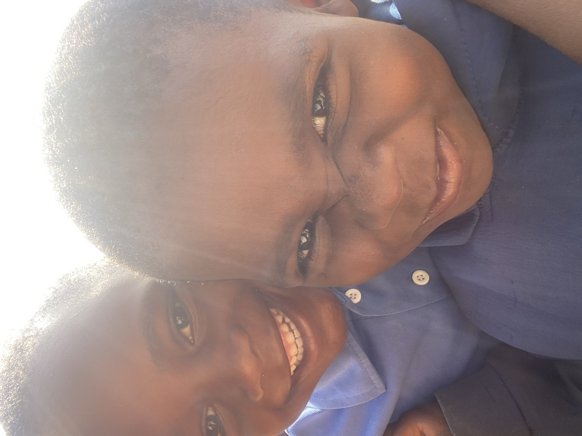 Two young Black males are pictured closely together, facing the camera. Both are wearing blue collared shirts. One has a broader face and is positioned closer to the top of the frame, with the other slightly below and to the side. They are both smiling. The environment is bright, potentially outdoors with strong sunlight causing overexposure on the left side of the image. The location is unspecified and not discernable.