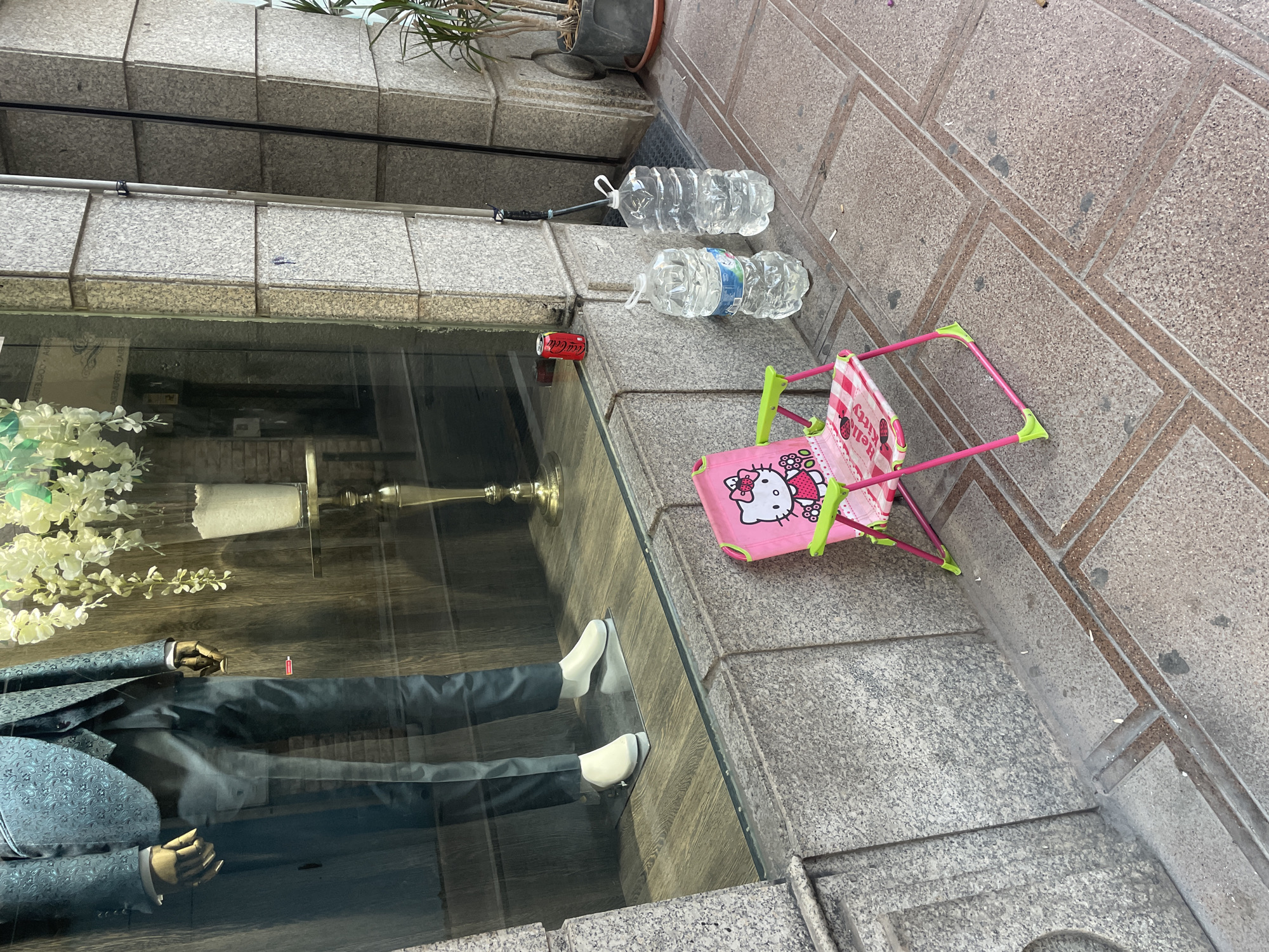 The image shows a street scene in La Línea De La Concepción, Spain. In the foreground, a child's pink folding chair with a Hello Kitty design sits on a sidewalk. Behind the chair, two large plastic bottles of water stand upright. To the left of the bottles, a red Coca-Cola can rests on a low stone ledge. Beyond the ledge, a shop window displays a mannequin dressed in a patterned teal suit, standing on a wooden floor. The building exterior is constructed of grey granite blocks.