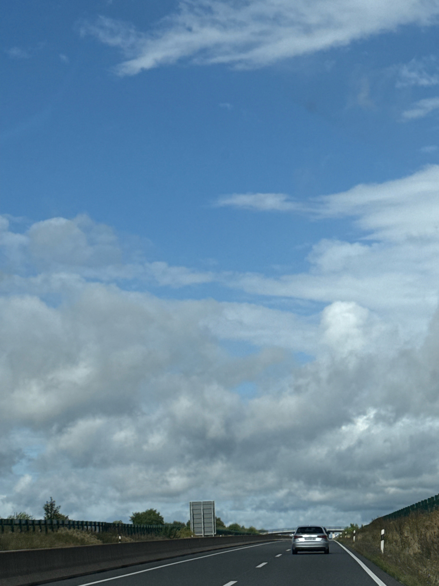 The image depicts a perspective from a moving vehicle on a highway, likely in or near the town of Bad Sachsa, Germany, given the contextual information. The sky is a prominent feature, characterized by a bright blue expanse with scattered white and gray clouds. The clouds vary in density, with some appearing wispy and others more substantial and textured.
In the foreground, the asphalt road is visible, with clear white lane markings. A concrete barrier runs along the left side of the road, separating it from the adjacent embankment. On the right side of the highway, a dry, grassy embankment rises, dotted with small shrubs and rocks. A dark green fence or railing runs along the edge of the embankment in places.
A silver car is traveling ahead on the highway. It is positioned in the right lane and is the primary subject on the road. In the distance, to the left of the road, a large rectangular sign structure is visible. Further back, there are trees and greenery. The overall environment suggests an open, possibly rural or semi-rural highway setting. The weather appears to be overcast with intermittent sun, typical of a cloudy day.