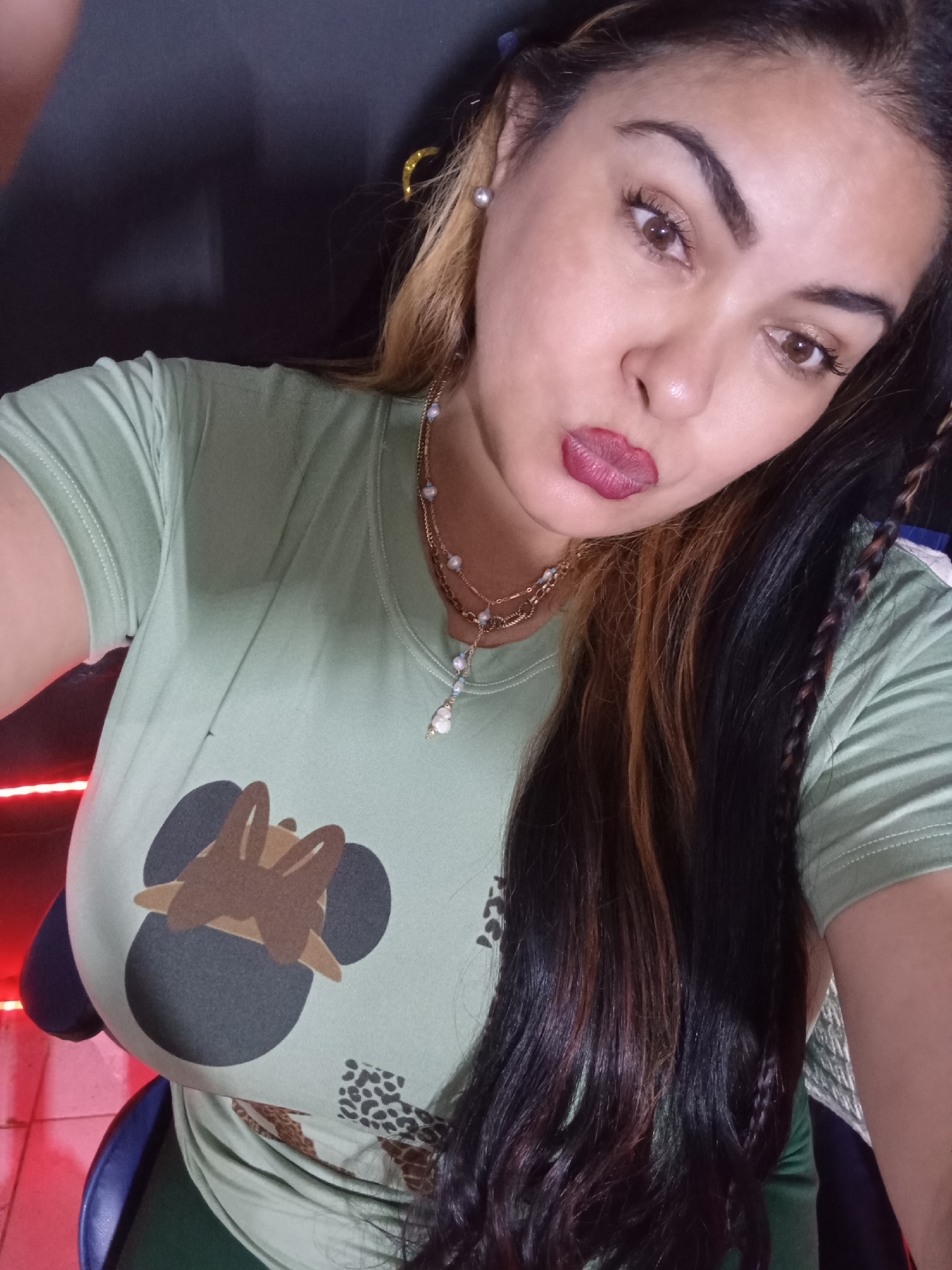 An adult female is depicted in a close-up, chest-up selfie taken in what appears to be an indoor setting in Juan Griego, Venezuela. Her gaze is directed slightly downwards and to the right, while her lips are pursed in a pout, accentuated by dark red lipstick. She has a medium complexion, dark brown eyes, and prominent dark eyebrows. Her long hair is predominantly dark brown with lighter, possibly blonde or reddish-brown, streaks visible on the left side. A small braid is visible over her right shoulder. She wears a light green short-sleeved t-shirt featuring a graphic design of a dark grey shape with brown, rabbit-like ears on the chest, and a subtle leopard print pattern below it. A multi-strand gold-toned necklace with small white and light blue beads and a pendant adorns her neck. A white pearl stud earring is visible in her left ear. The background is dark and out of focus, with a distinct red light source visible on the lower left, illuminating a partial blue object, possibly a chair. The floor in the lower left corner appears to be light-colored.
