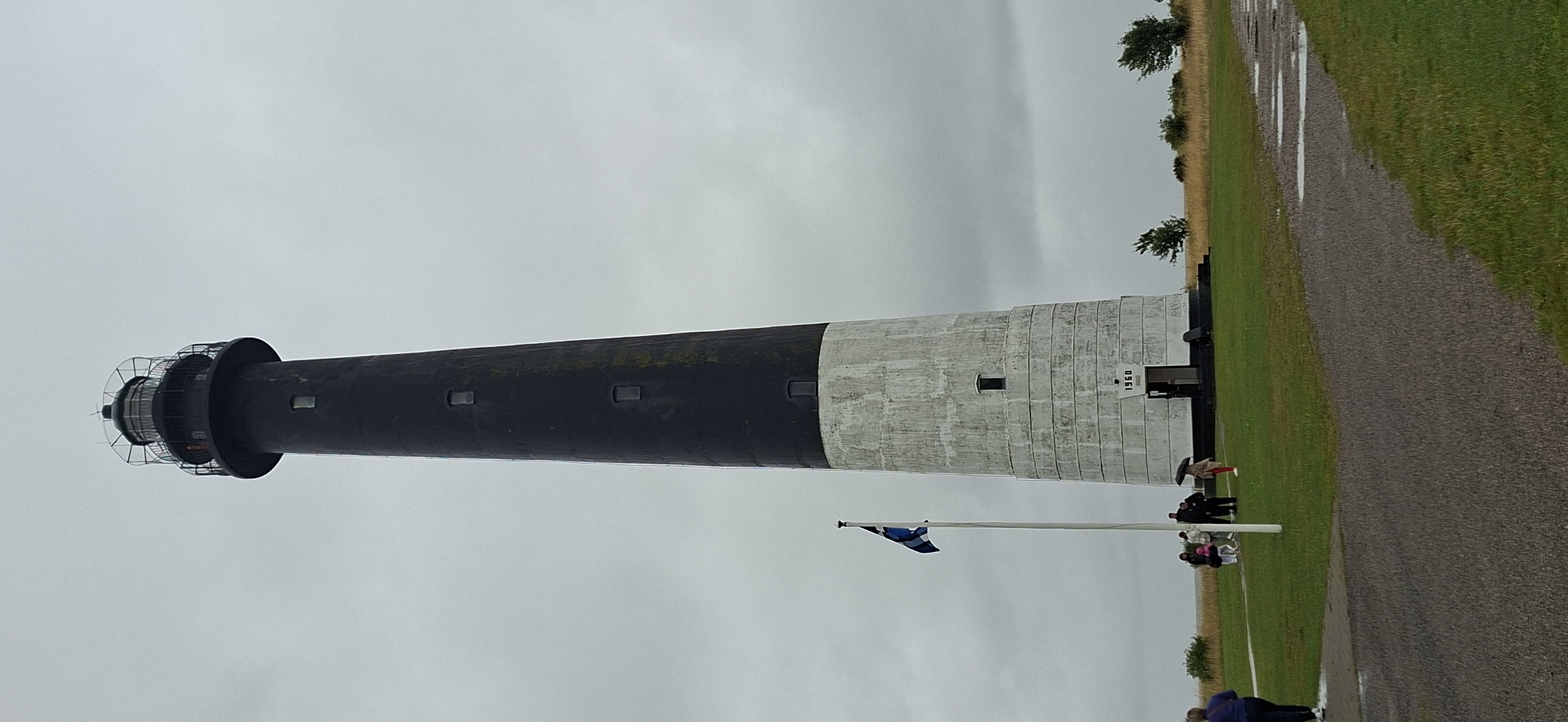 A tall, tapering lighthouse with a black top section and a white, weathered lower section stands against a cloudy sky. The lighthouse has several small, rectangular windows and a lantern room at its peak. A flag pole with a blue and white flag is positioned to the left of the lighthouse. Several people, some holding umbrellas, are gathered near the base of the lighthouse. The ground in front is covered with green grass and a paved path. The lighthouse is labeled with the year "1960". The scene is the Sääre lighthouse on the Sõrve Peninsula in Estonia.