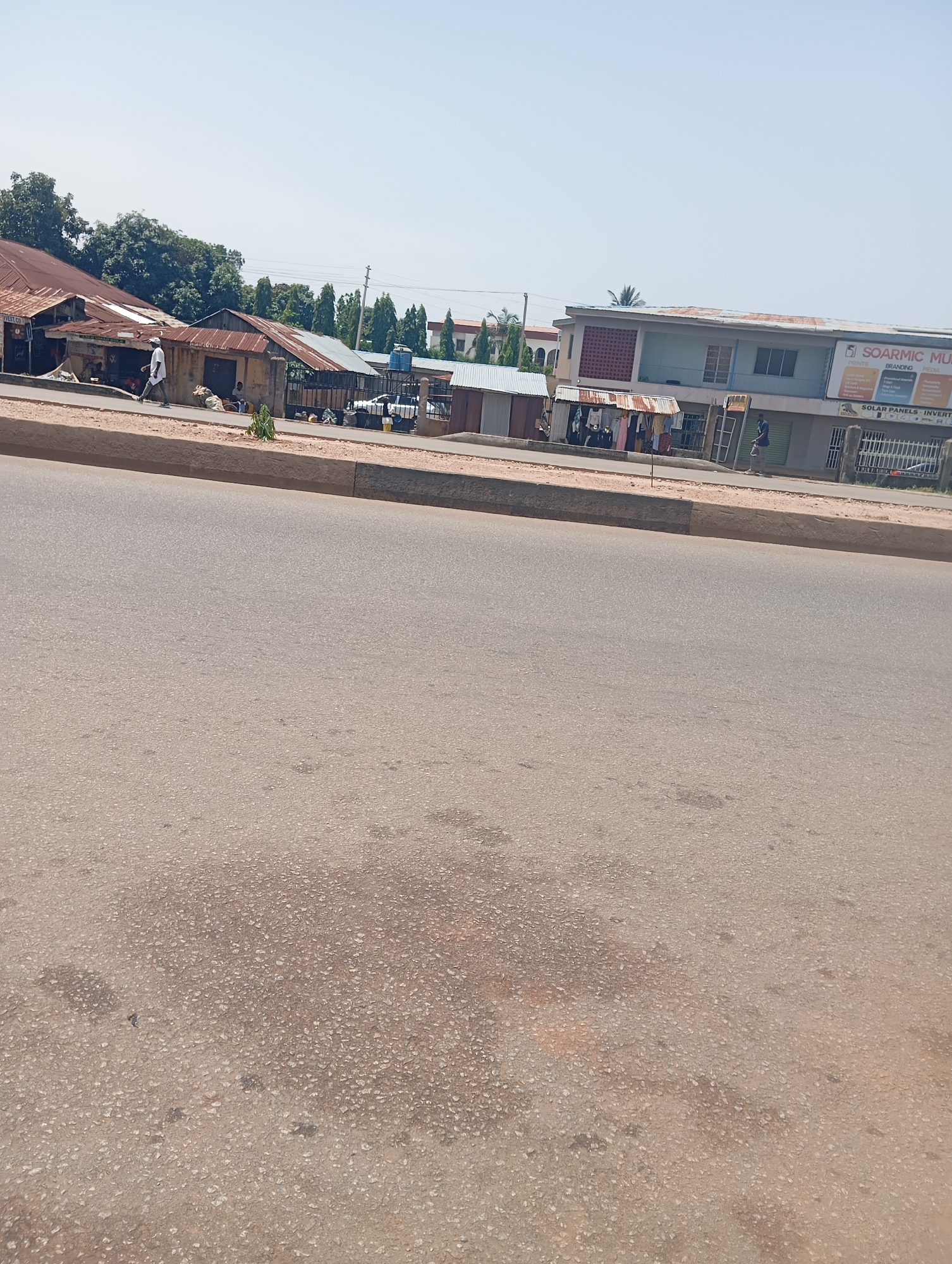 The image captures a street scene in Jos, Nigeria, under a bright, clear sky, suggesting midday. A paved road occupies the foreground, with a raised concrete curb separating it from the sidewalk and buildings beyond. On the sidewalk, several small businesses are visible. To the left, a row of simple structures with corrugated metal roofs appear to be shops, with goods displayed outside. A person in a white shirt and dark pants is walking along the sidewalk. Further to the right, a more substantial two-story building stands, with shops on the ground floor. A sign on this building reads "SOARMC MU" and below that, "SOLAR PANELS - INVERT". Another sign on the building indicates services like "BRANDING" and "MEDIA." People are seen on the sidewalk, some standing near the shops, and others potentially interacting with the businesses. A white vehicle is parked near the buildings. The overall impression is a busy commercial area in a Nigerian city, with evidence of trade and everyday life. The weather appears to be warm and sunny.