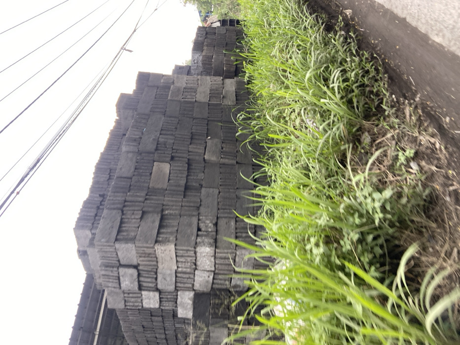 A stack of gray concrete blocks are piled high against a grassy embankment. Green grass and weeds grow along the embankment. The blocks are stacked neatly in a row and there are several power lines running overhead. The scene is located in the Kecamatan Margaasih region of Indonesia, at latitude -6.961 and longitude 107.536.