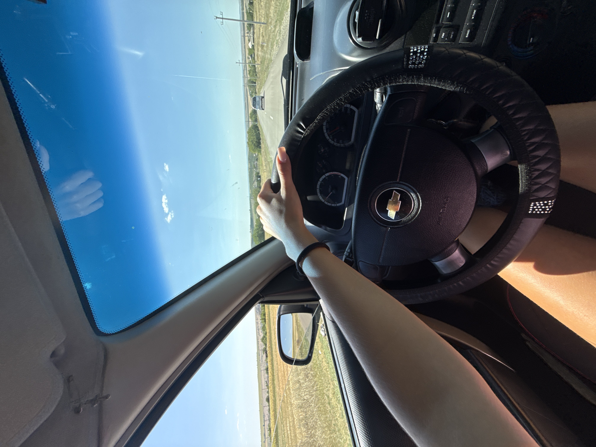 This is a first-person perspective shot from the driver's seat of a car traveling down a road in Basarabeasca, Moldova. The driver's hands are visible on the steering wheel, which is covered in a black, diamond-patterned material with rhinestone accents. The dashboard and part of the car's interior are visible. Through the windshield, a two-lane road stretches into the distance, with a white car ahead. The landscape on either side of the road consists of dry, golden fields, scattered trees, and rural buildings, suggesting a sunny, possibly late summer or early autumn day. The sky is clear blue with a few wispy clouds. The side mirror reflects the road and the rural scenery.