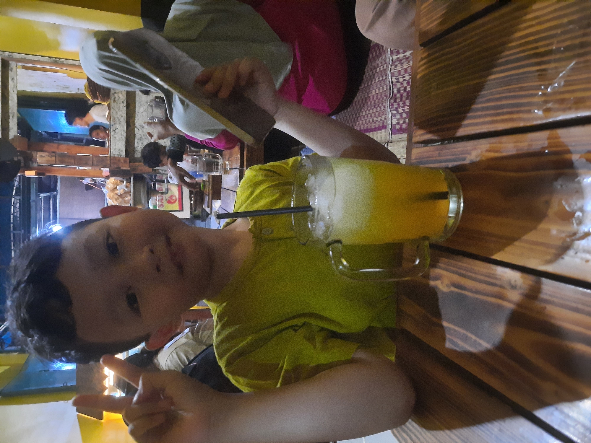 The image depicts a young boy in a brightly colored yellow shirt, posing for the camera with a peace sign. He is seated at a wooden table, with a glass of orange juice and ice in front of him. Behind the boy, other people are visible, some blurred in the background. One person, wearing a light-colored headscarf and a pink garment, is partially visible, holding what appears to be a book or menu. The setting appears to be an indoor eatery with a rustic, wooden interior. The lighting suggests an evening or dimly lit environment. The overall atmosphere is casual and social. This scene is likely taking place in an informal dining establishment in North Jakarta, Indonesia, characterized by its local ambiance and communal seating arrangements.