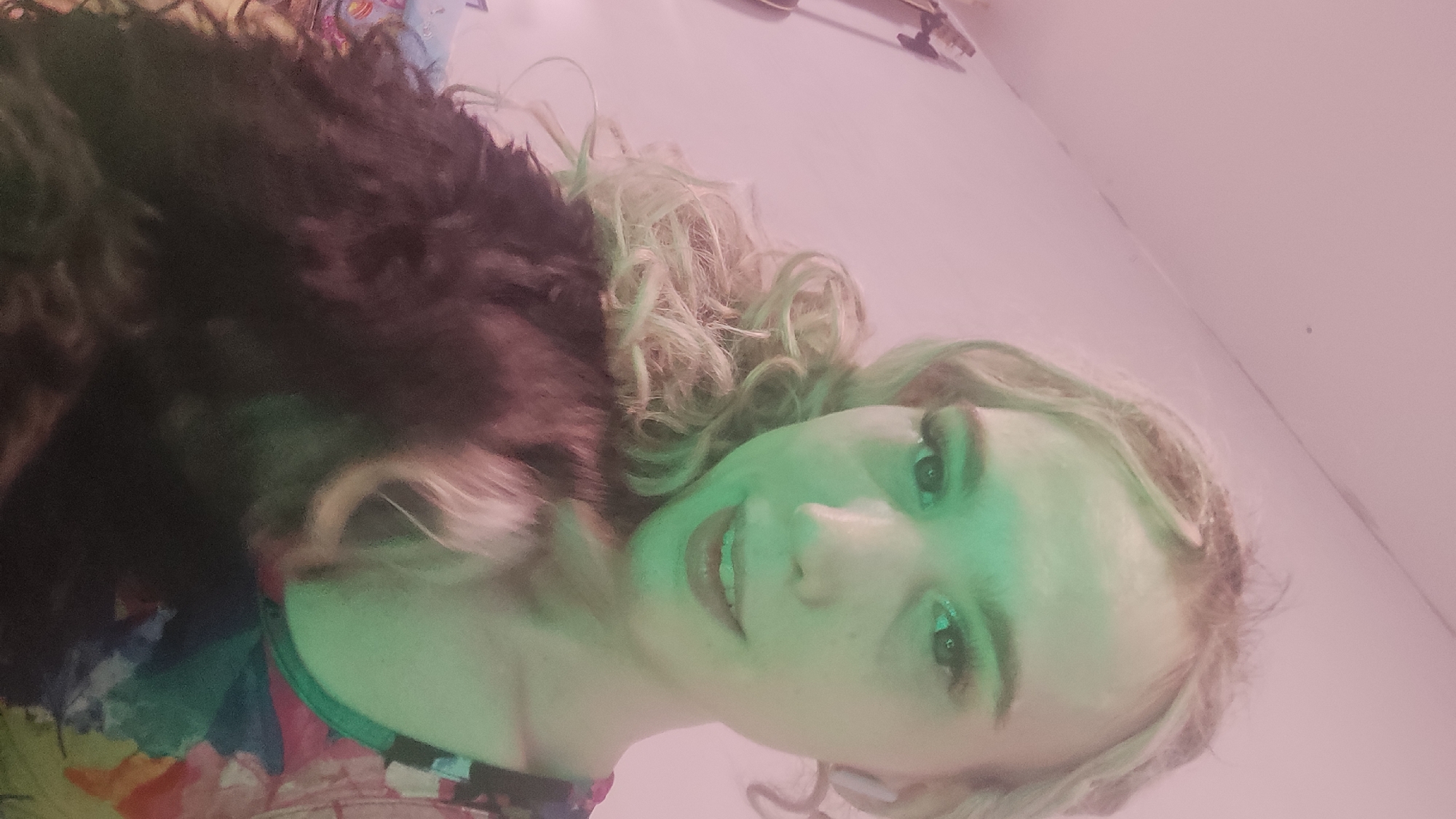 A selfie shot depicts a person with fair skin and blond, curly hair. The person is partially obscured by a dark, furry object, possibly a coat or animal. The background appears to be a pale wall. The location is unspecified. The lighting is uneven.
