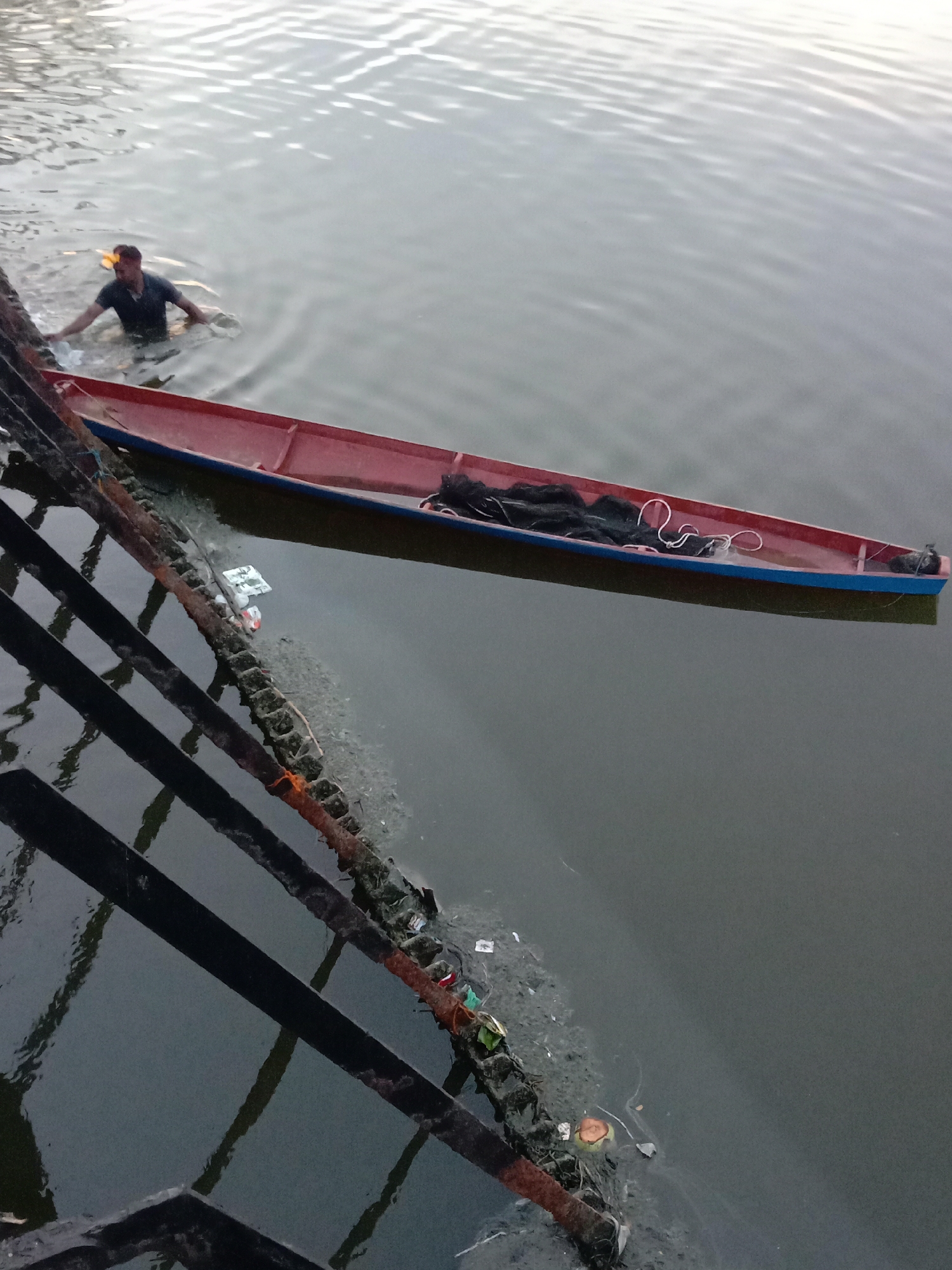 The image depicts a man waist-deep in a body of water, likely a river or canal, near a bridge. A narrow boat, painted red with a blue trim, floats beside him. A fishing net and white rope are inside the boat. The water is murky, and debris is visible along the bridge's structure and in the water. The image appears to be taken from the bridge looking down. Considering the location of Lhokseumawe, Indonesia, it is likely a local waterway. The man appears to be interacting with the boat.